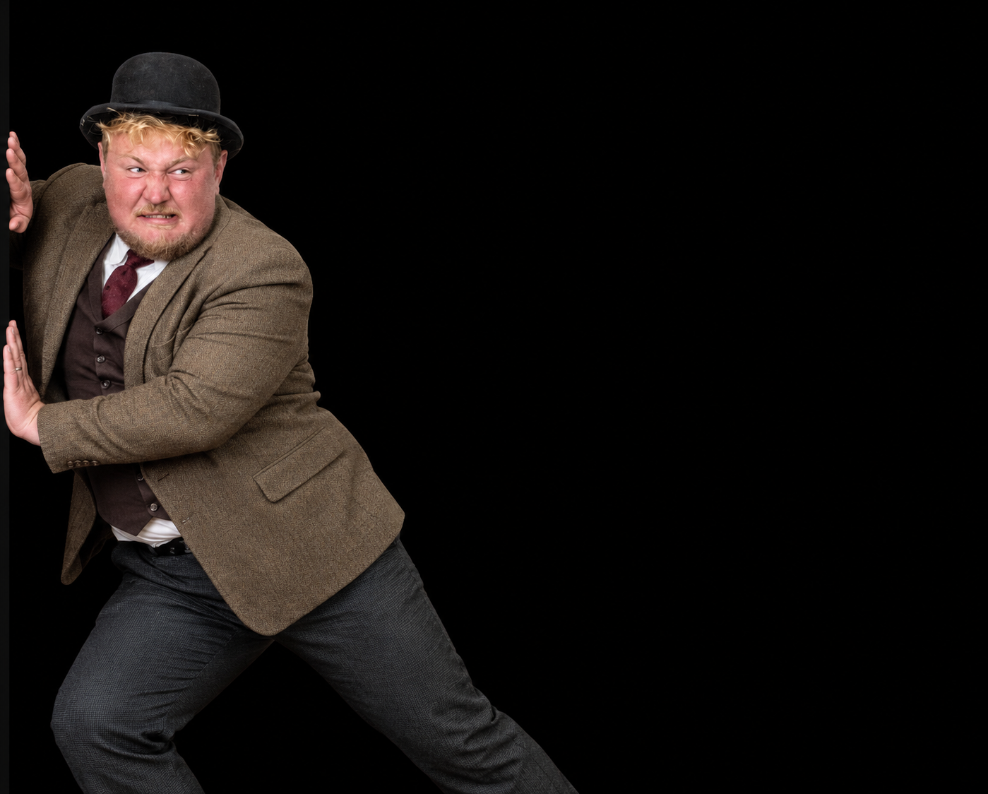 A man dressed as a magician or performer, wearing a brown blazer, gray pants, a white shirt, a dark vest, and a black bowler hat, poses with a playful and intense facial expression against a black background.