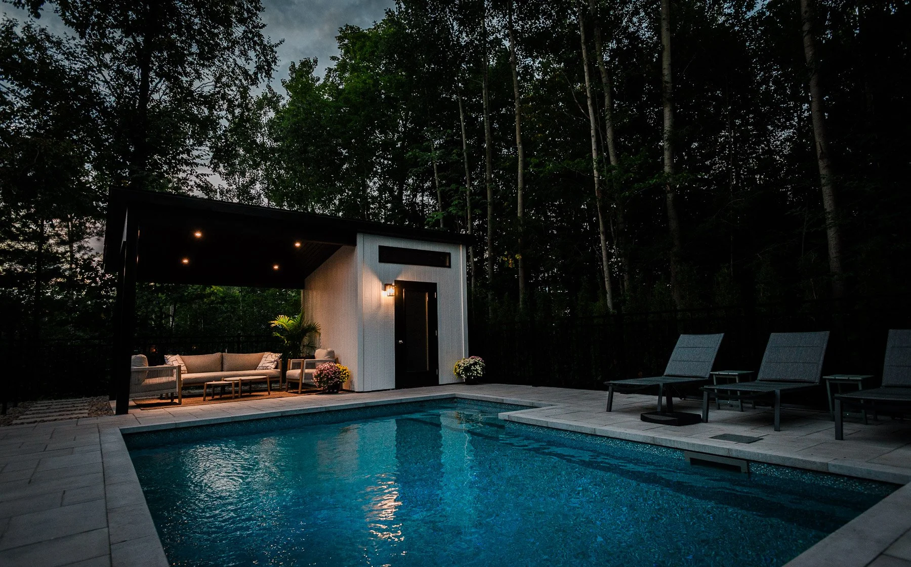 Piscine éclairée en soirée avec mobilier extérieur, petites fleurs et maison moderne en arrière-plan, entourée d'arbres.