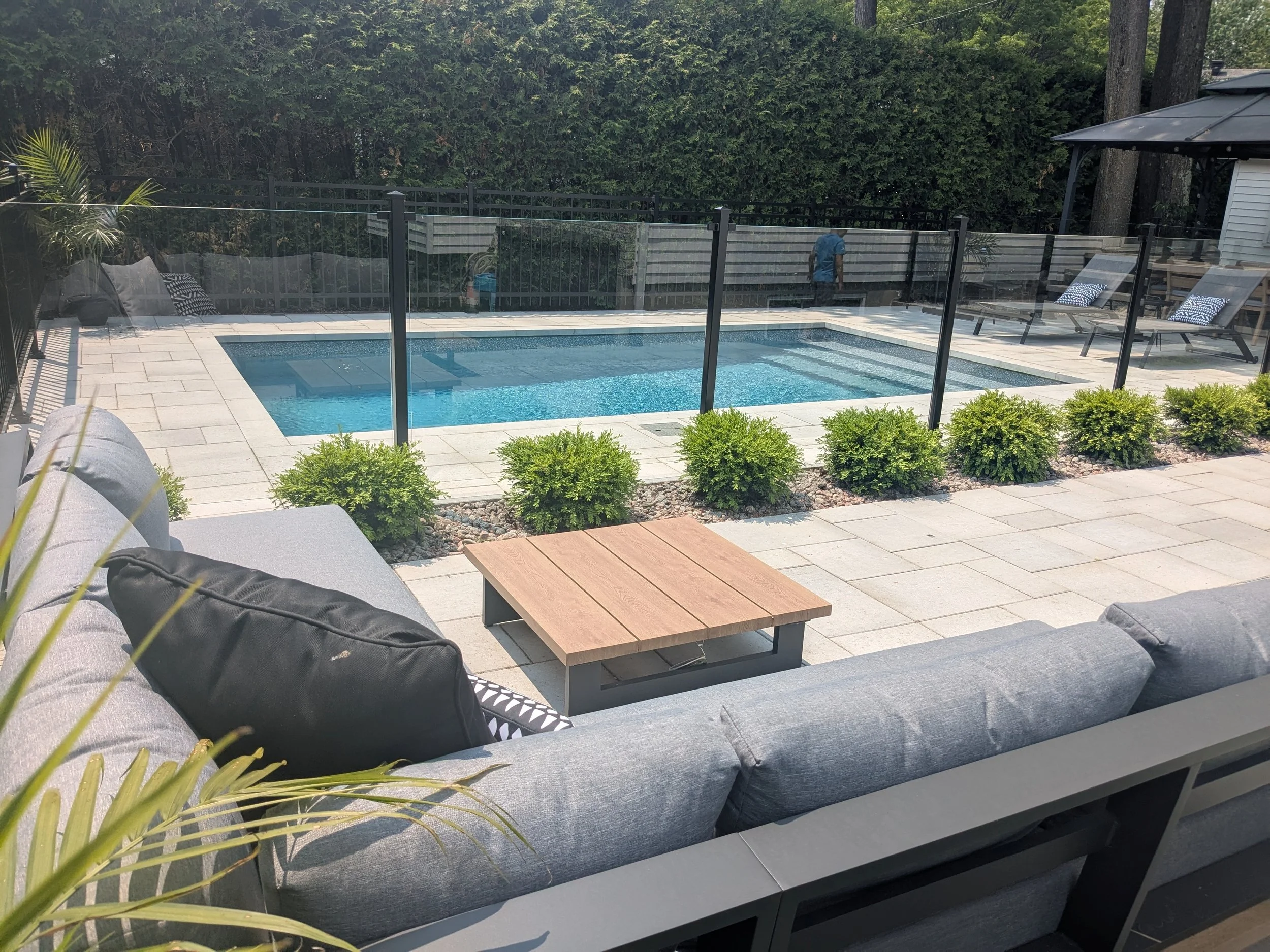 Vue d'une terrasse avec un spa ou piscine, des fauteuils, des coussins et un parasol, entourée de haies et d'arbres.