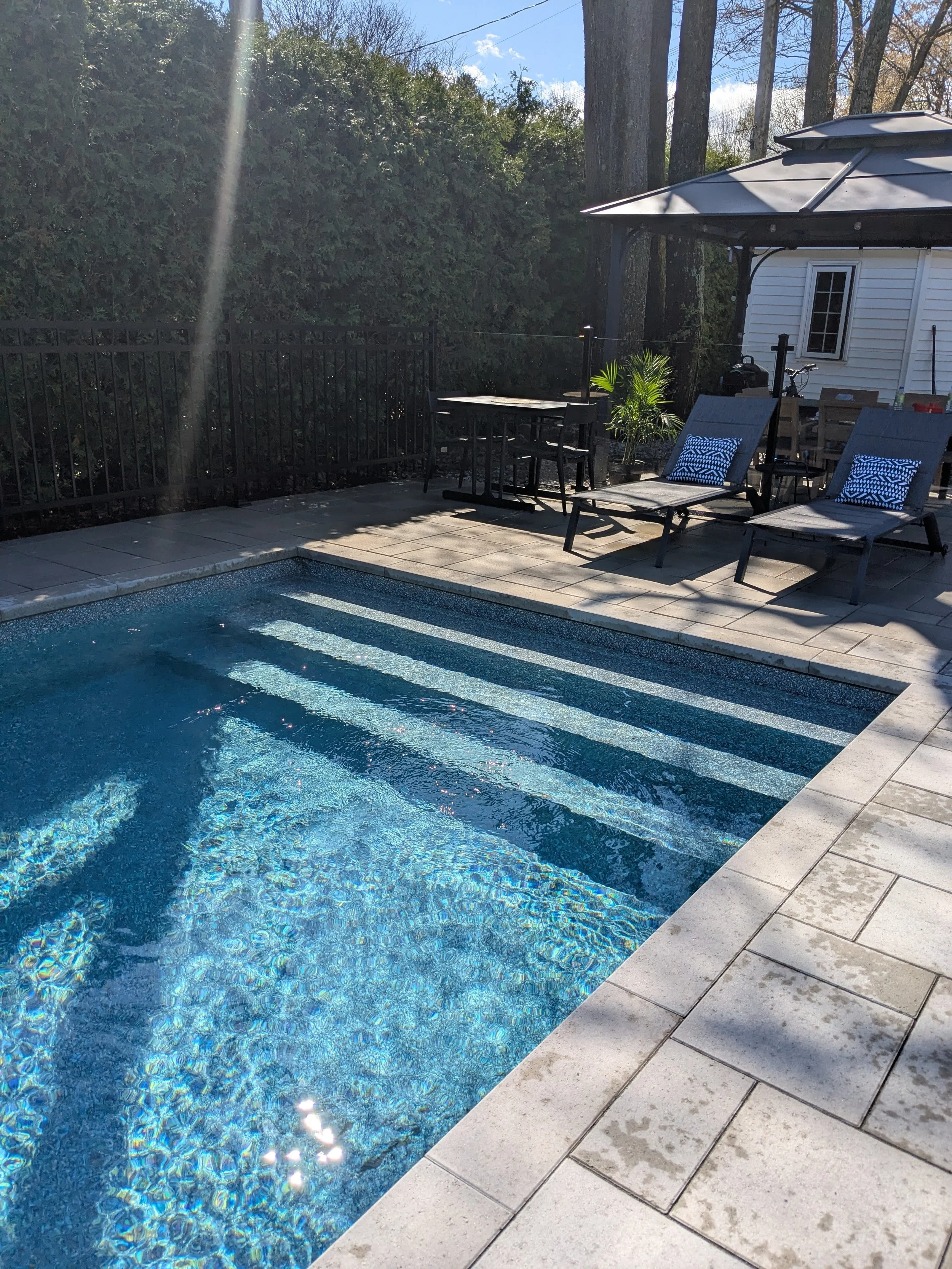 Piscine extérieure à débordement en béton entourée d'une terrasse en carreaux, avec deux chaises longues, un parasol, et un petit bâtiment en arrière-plan avec des arbres et un ciel clair.