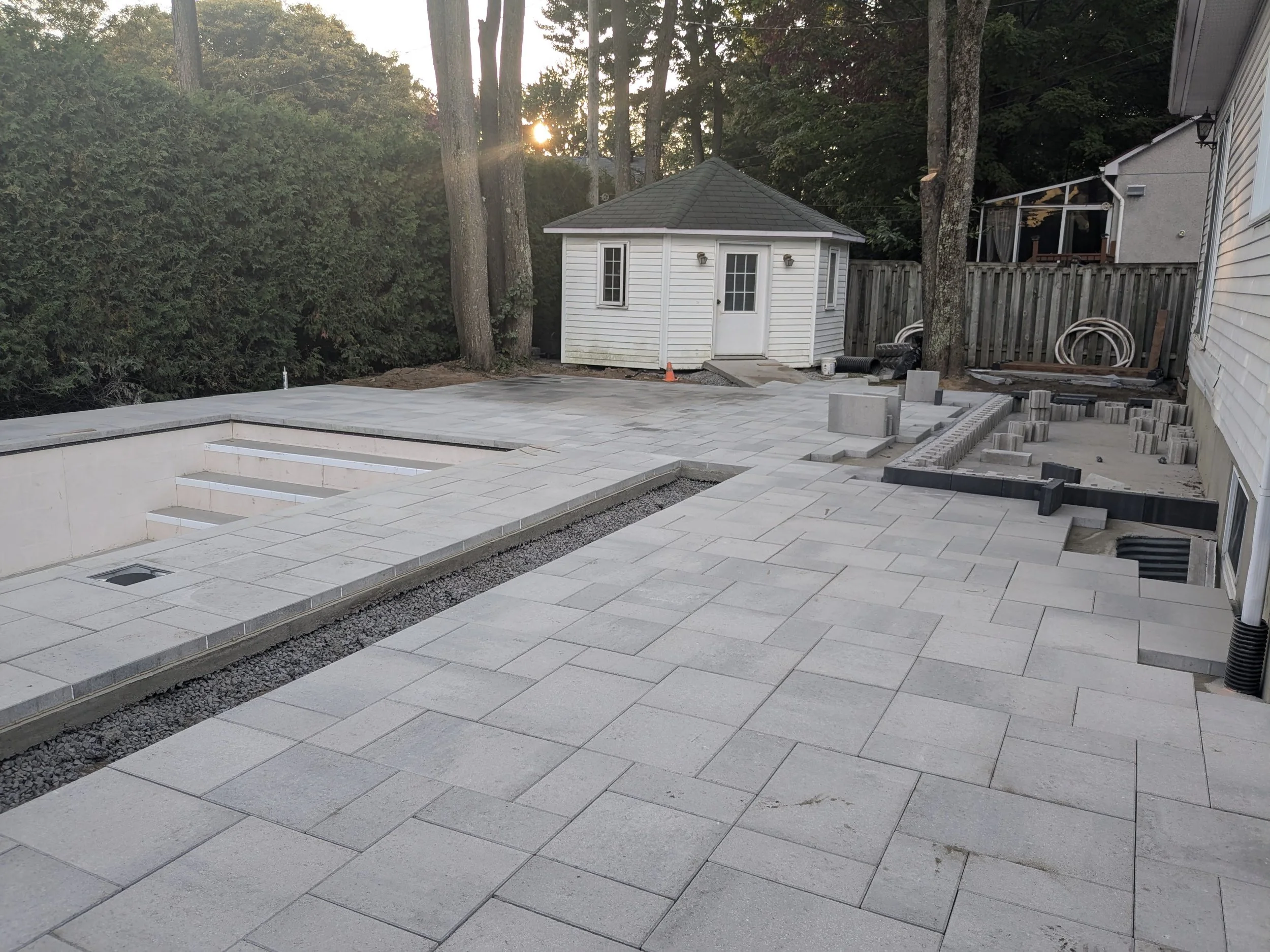 Un patio en cours de rénovation avec pavés en cours de pose, une petite cabane blanche avec porte en verre, arbres et une maison en arrière-plan, le soleil couchant.