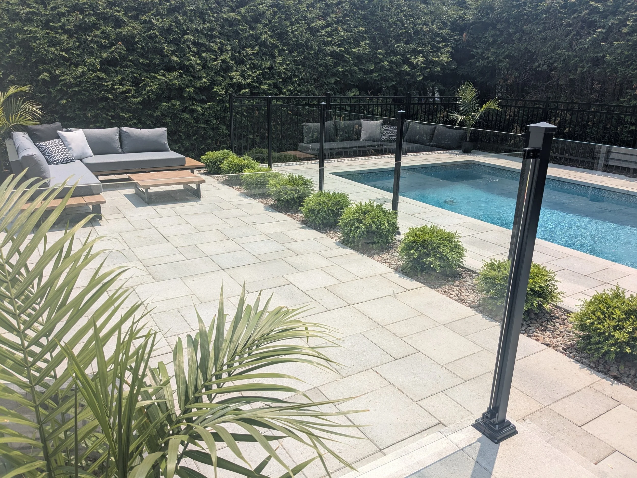 Une terrasse avec un canapé gris, une petite table en bois, des plantes en pot, et une piscine entourée d'une clôture en métal, avec un fond de haies vertes.