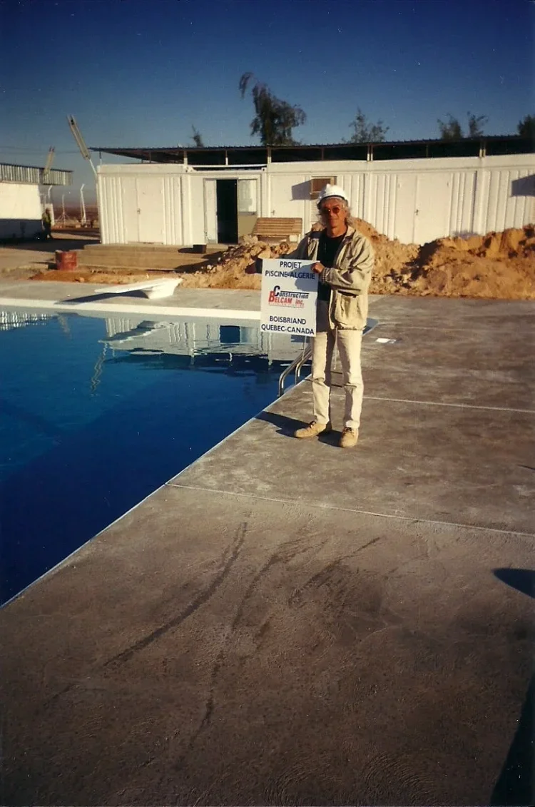 Une personne posant à côté d'une piscine en construction, tenant une affiche mentionnant le projet de construction d'une piscine en Algérie, au Québec, Canada, avec un bâtiment en arrière-plan et un ciel nocturne.