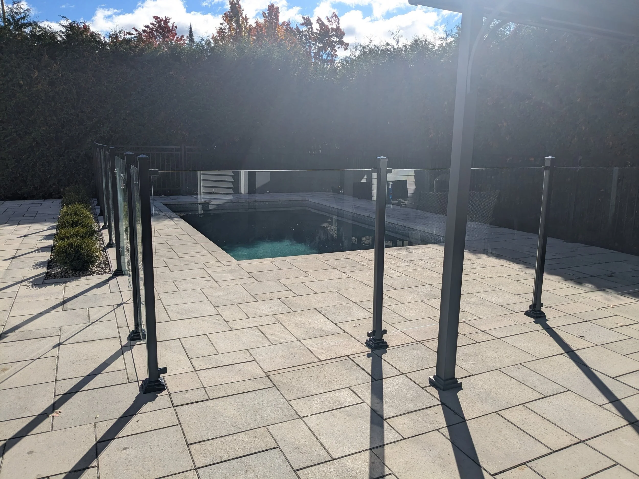 Une piscine extérieure entourée d'une terrasse en pavés et d'une clôture en verre, avec des arbustes plantés le long du pourtour. En arrière-plan, des arbres aux feuillages d'automne et un ciel partiellement nuageux.