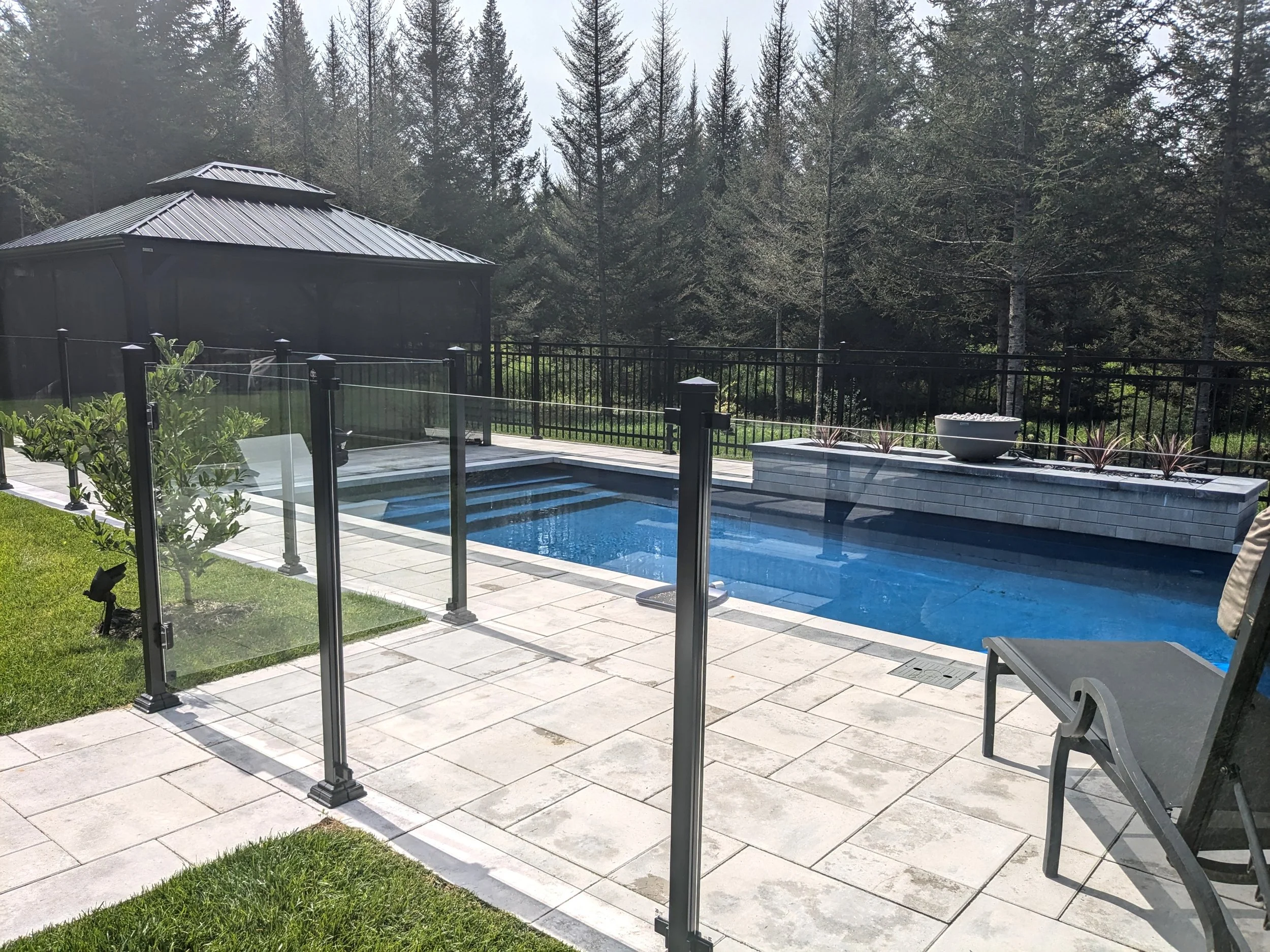 Une piscine extérieure entourée d'une terrasse en pierre, avec un garde-corps en verre noir, un cadre de soleil, des chaises longues en plein air, des plantes en pot et une forêt d'arbres derrière, sous un ciel nuageux.
