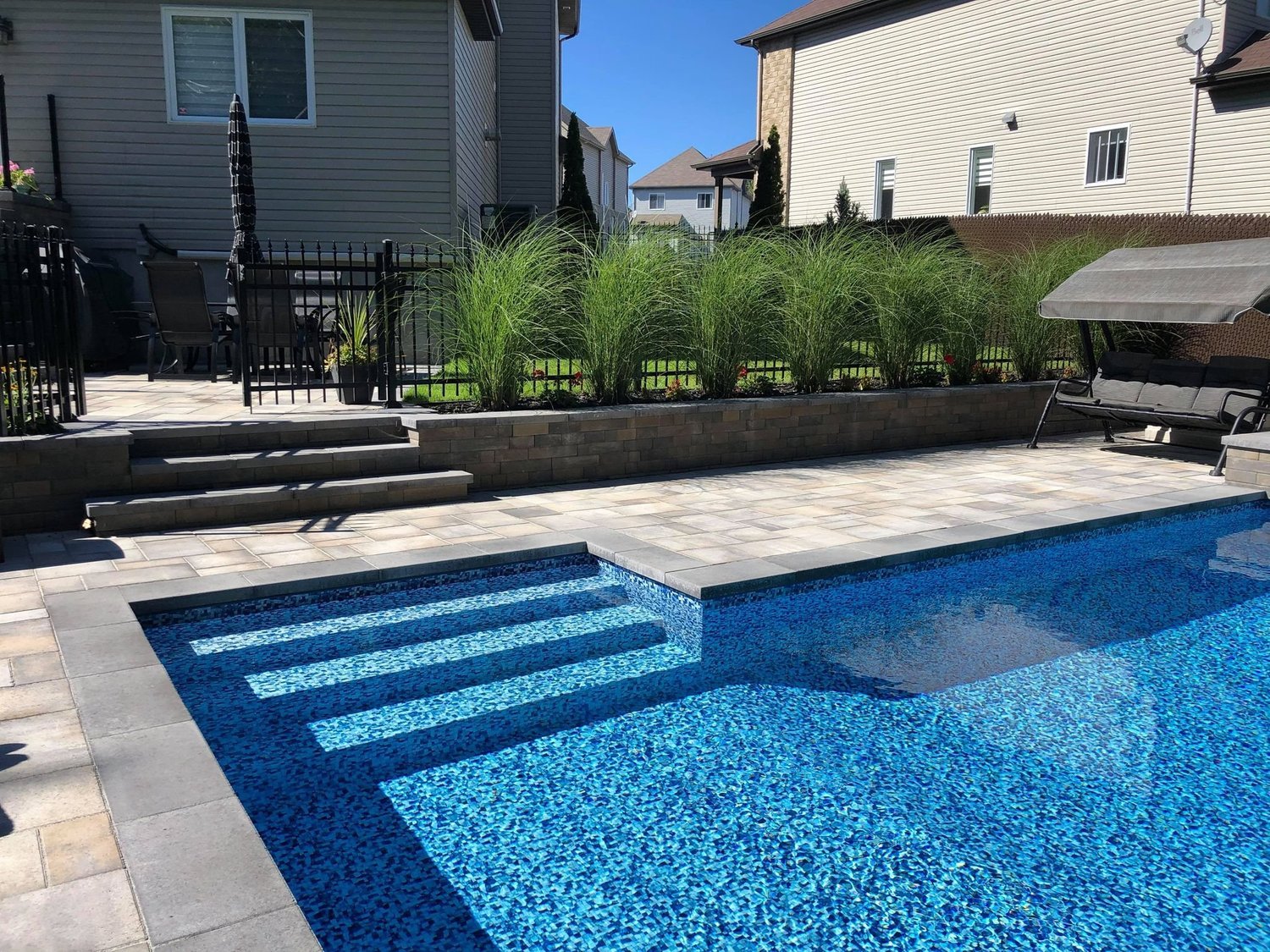 Piscine extérieure avec des escaliers en mosaïque bleue, terrasse en pierre, buisson d'herbes hautes et mobilier de jardin dans un arrière-cour résidentielle
