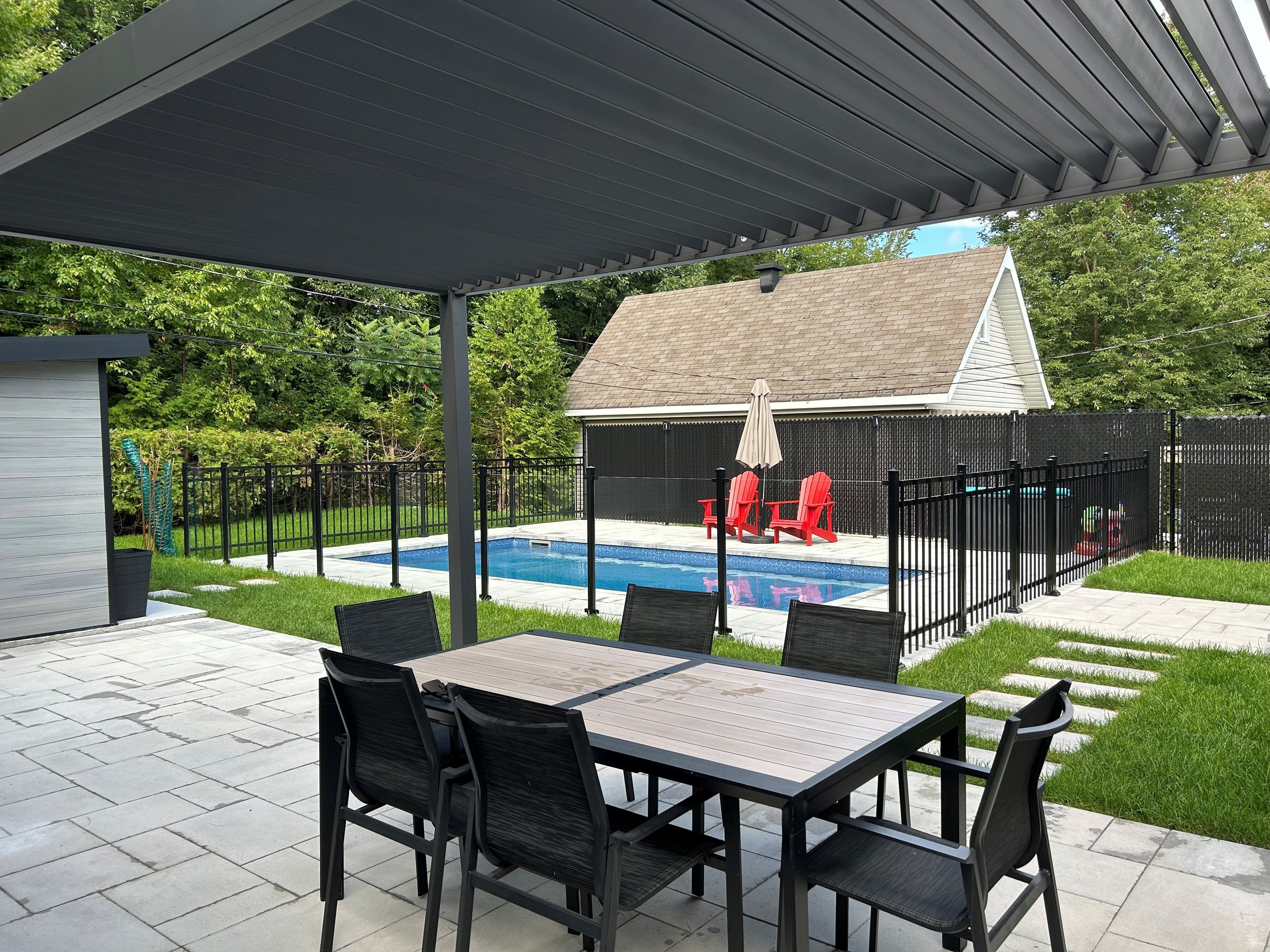 Terrasse avec table et chaises en plastique noir, vue sur un piscine, un espace avec des chaises rouges, une clôture en métal noir et un mur en arrière-plan, avec un parasol plié et un ciel partiellement visible.