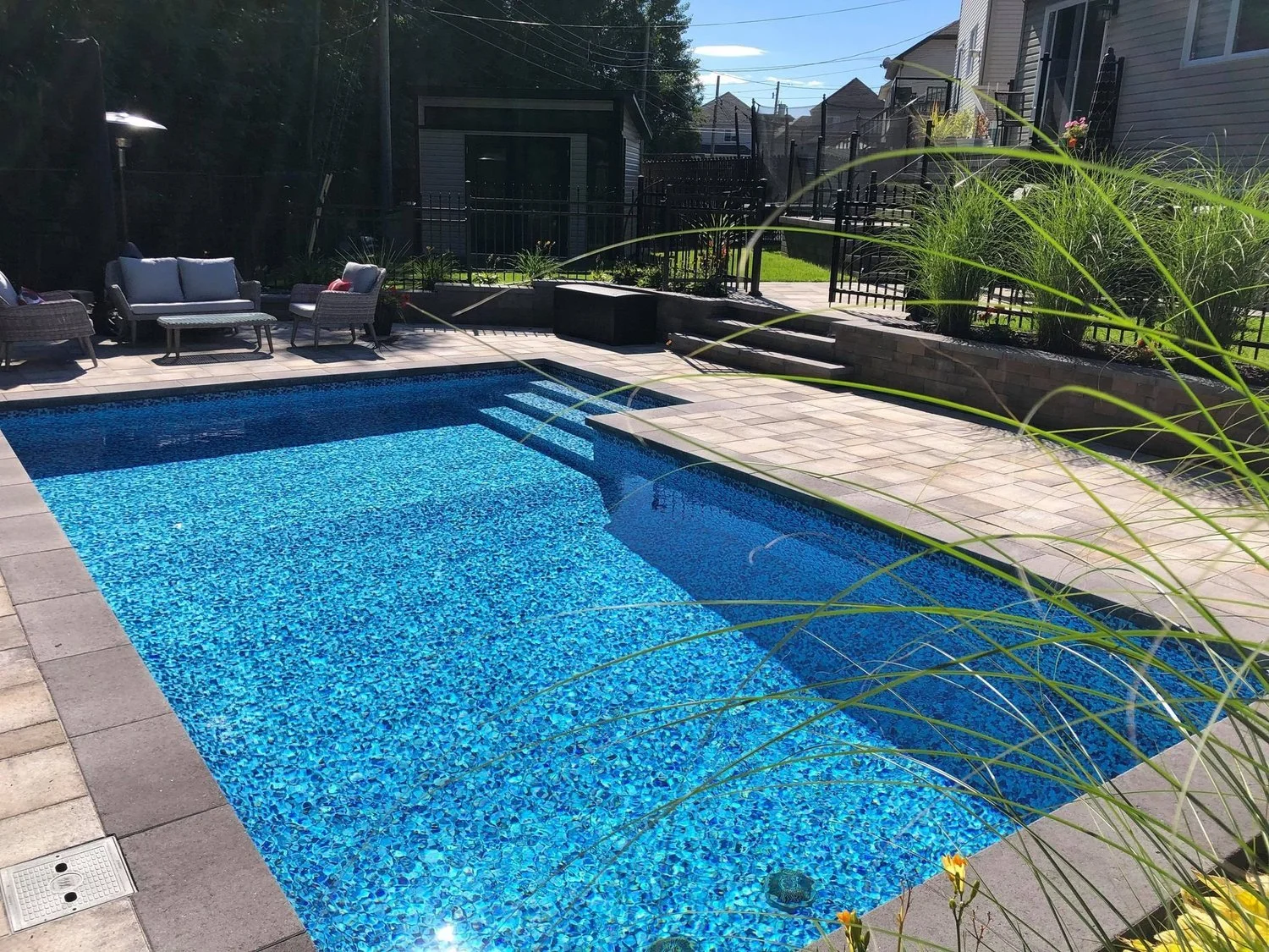 Piscine creusée en béton, avec des eaux bleues, entourée d'une terrasse en pavés et un espace avec des fauteuils en rotin pour se détendre, dans un jardin privé en arrière-cour. Des herbes ornementales en premier plan et une clôture en métal entouren