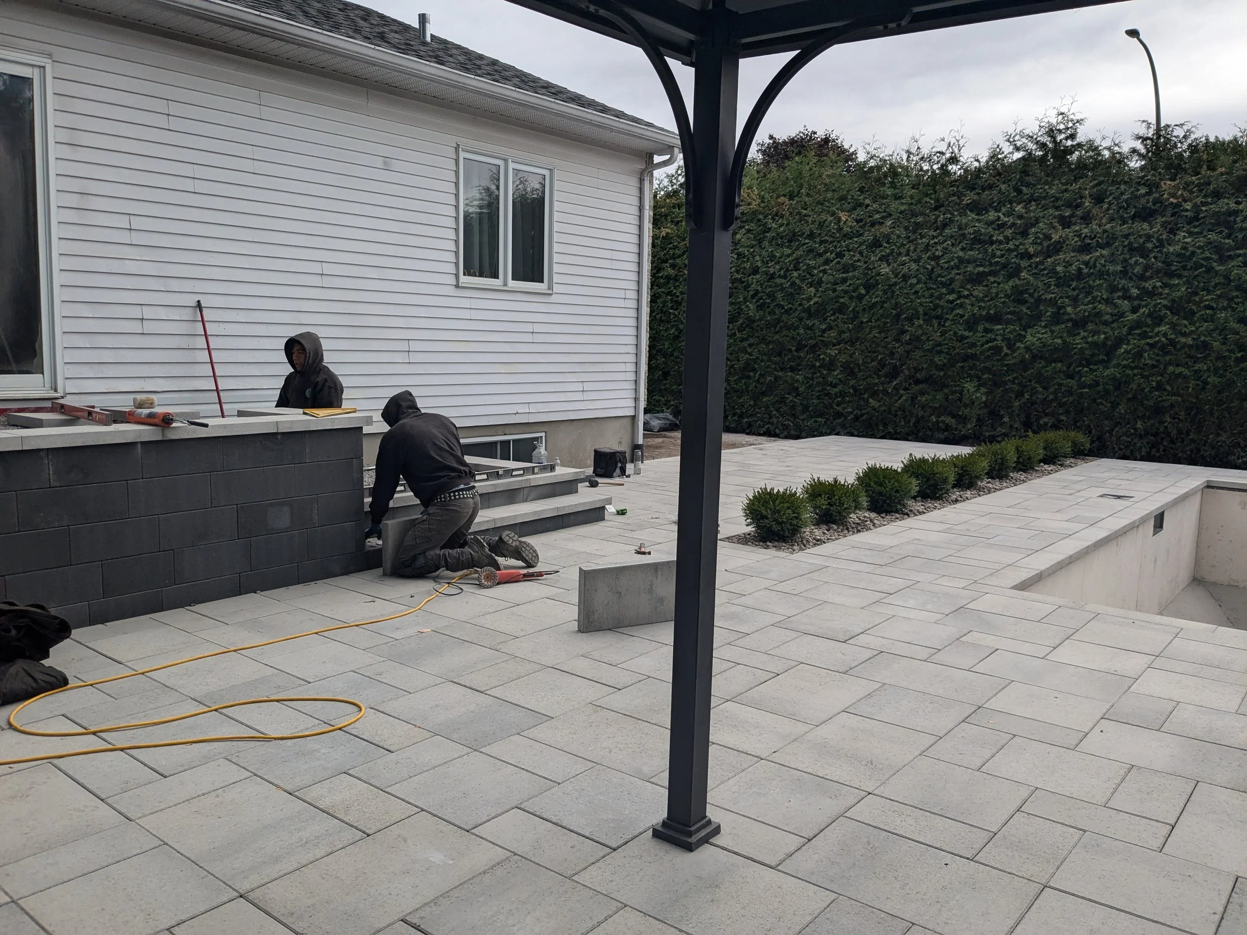 Deux personnes travaillent sur la terrasse d'une maison avec des outils et matériaux de construction, en train de poser ou de réparer des escaliers en pierre.