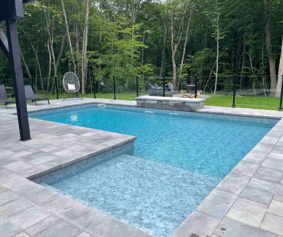 Piscine moderne entourée de pavés, avec des chaises longues, un fauteuil suspendu, et une zone de feu de camp, dans un cadre naturel forestier.