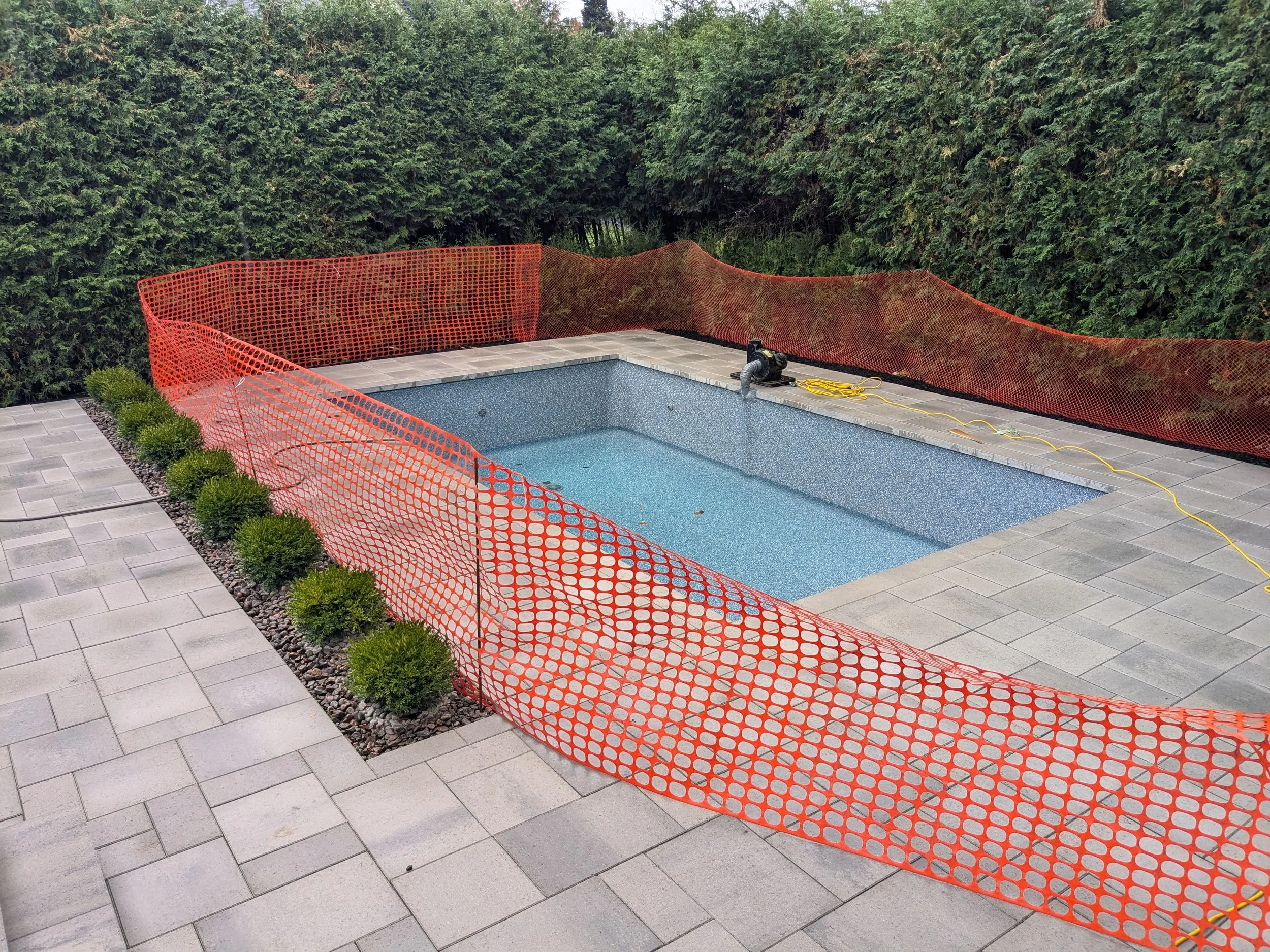 Une piscine vide entourée d'un trottoir en pavés et clôturée par une barrière orange de sécurité. Des plantations verdoyantes bordent un côté de la piscine.