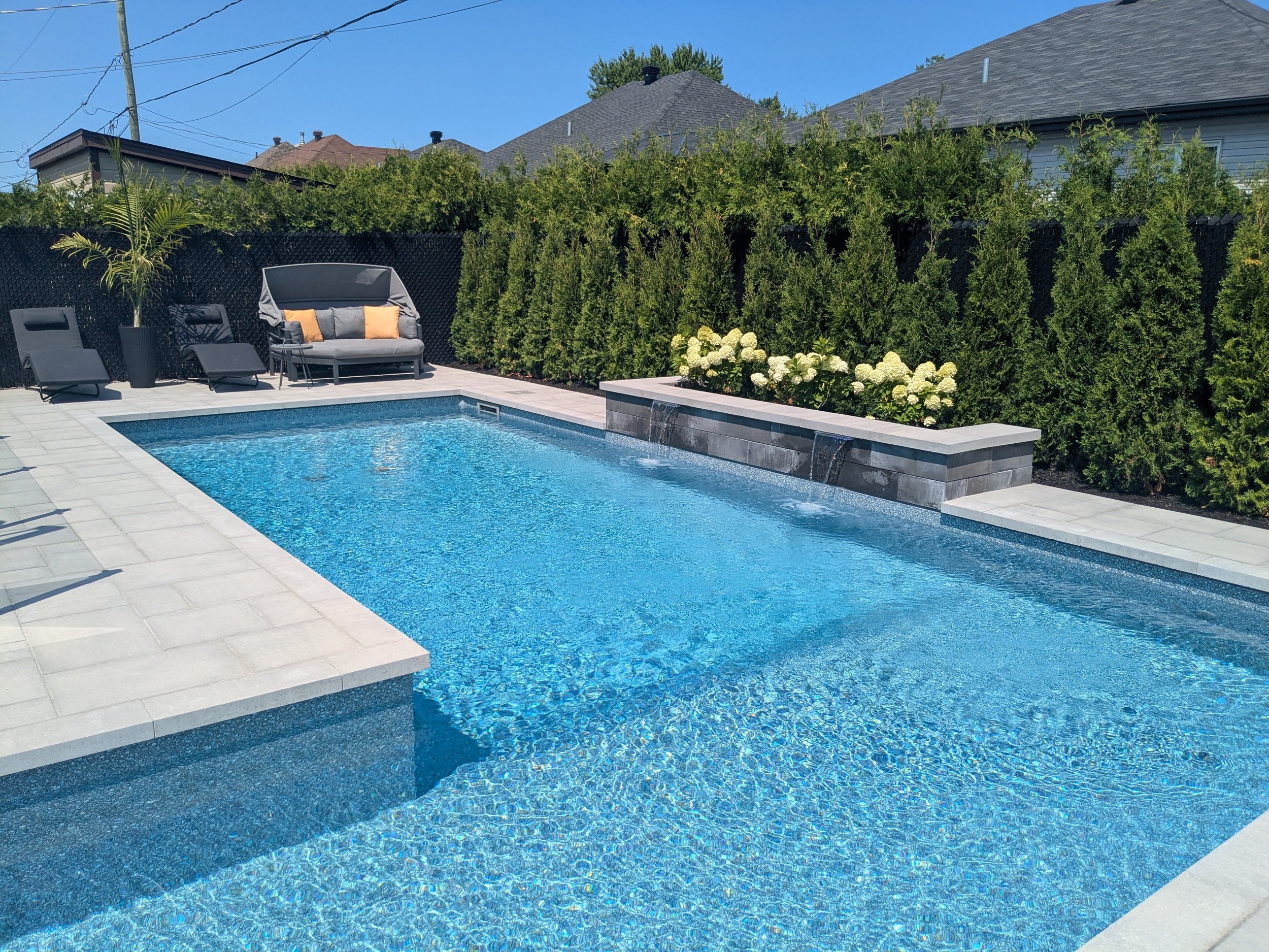 Une piscine avec une terrasse en pierre, entourée de haies et d'arbres, avec des chaises longues et un canapé de jardin.