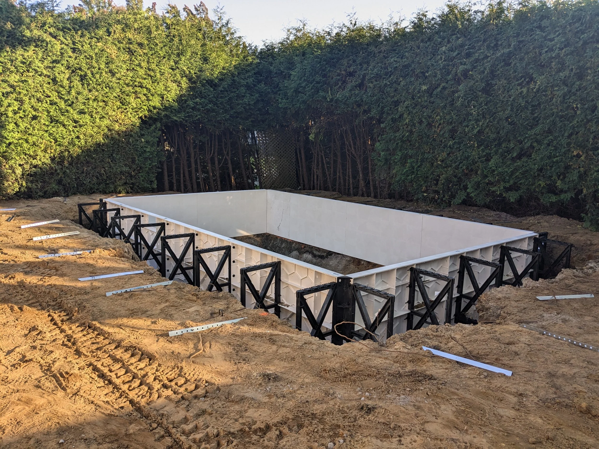 Une piscine vide en cours de construction, entourée de terre et de systèmes de support, avec des haies d'arbustes verts en arrière-plan.