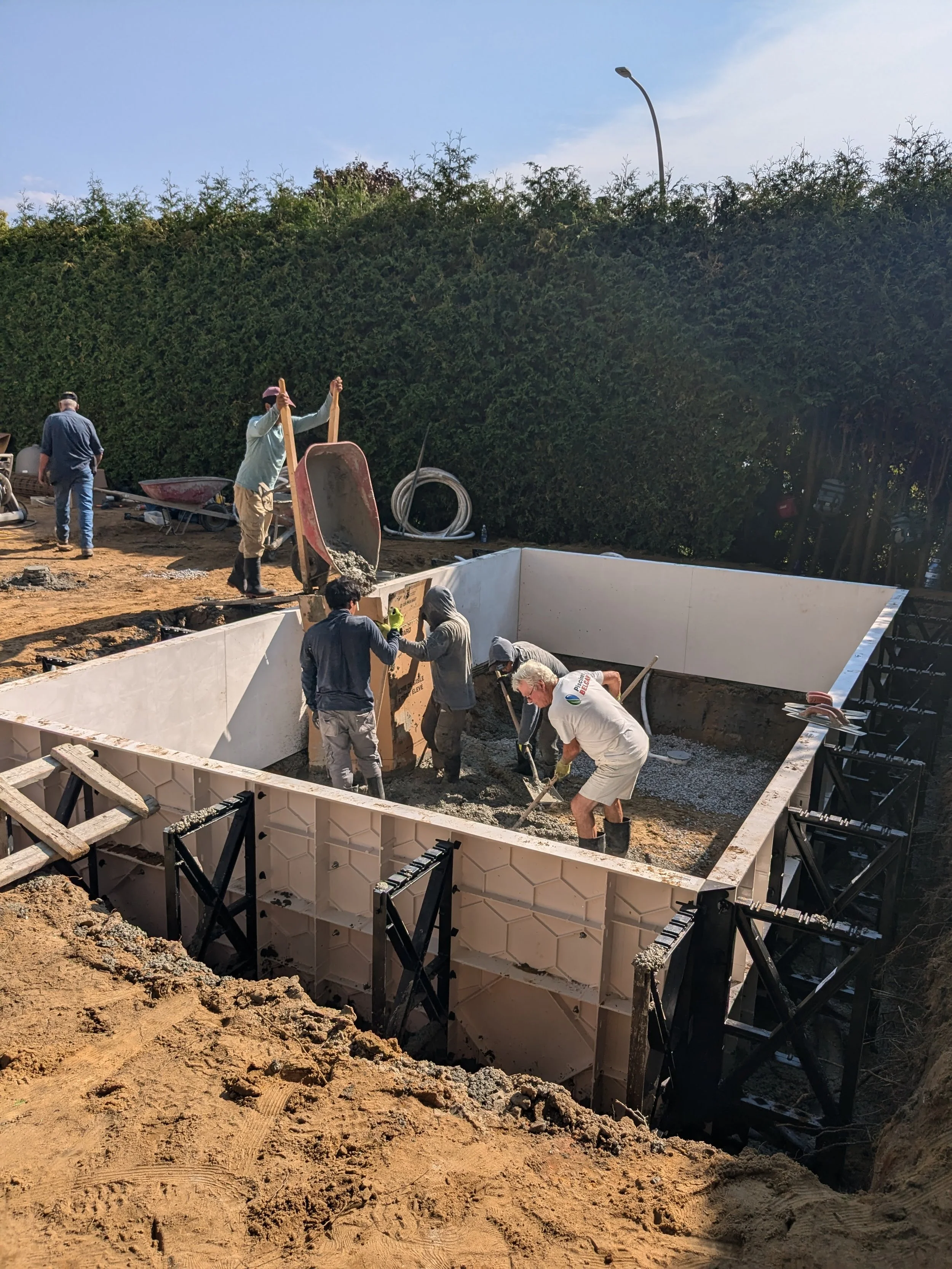 Chantiers de construction avec plusieurs ouvriers travaillant dans un trou profond, utilisant des outils pour le béton. Des brouettes, tuyaux et structures en métal sont visibles, avec un grand arbuste en arrière-plan et un ciel clair.