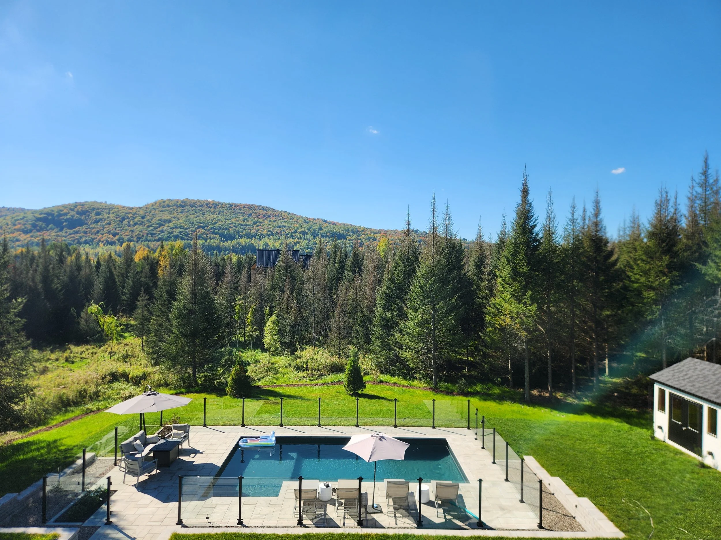 Piscine extérieure avec meubles et parasols, terrasse et jardin vert, entouré d'arbres et de montagnes sous un ciel bleu.