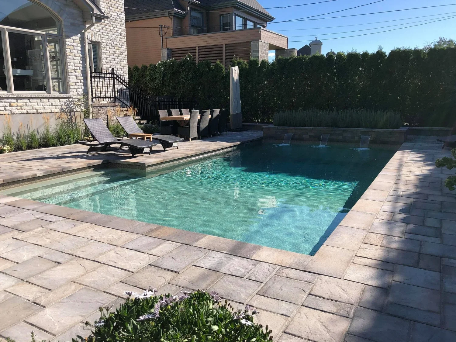 Piscine dans un patio en pierre avec chaises longues, un parasol fermé, une table en bois avec des chaises, et un mur végétal en arrière-plan.