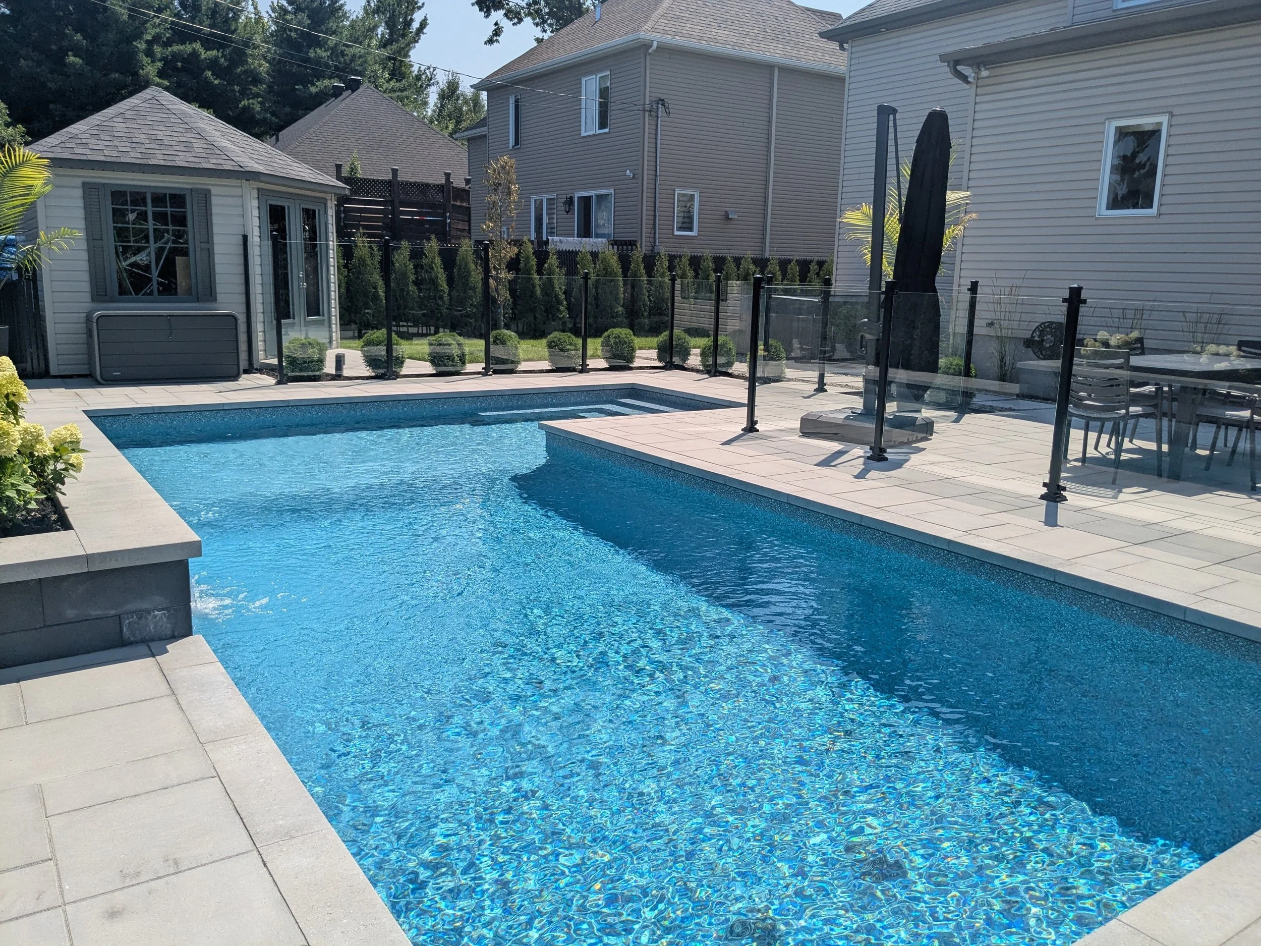 Une piscine extérieure entourée de dalles en pierre, avec une clôture en verre. Il y a des bâtiments résidentiels en arrière-plan, avec des arbres et un ciel ensoleillé.