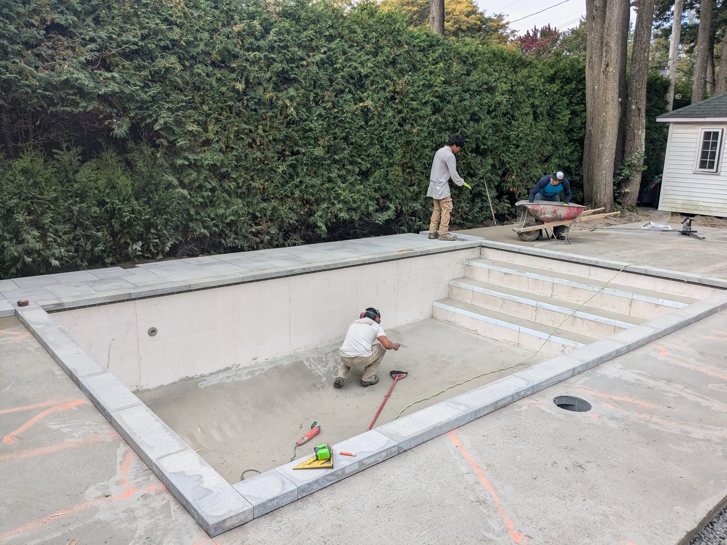 Deux hommes travaillent à la construction d'une piscine en béton, entourée d'une structure en pierre avec des escaliers, dans une cour arrière avec un grand mur d'arbustes et des arbres en arrière-plan.