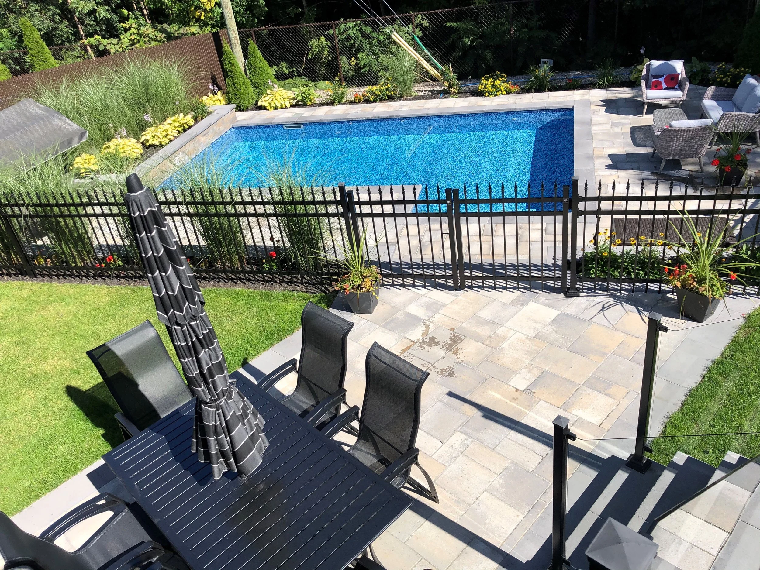Une piscine rectangulaire bleue entourée d'une terrasse en pierre, avec des meubles d'extérieur en rotin et des plantes ornementales, clôturée par une barrière noire.