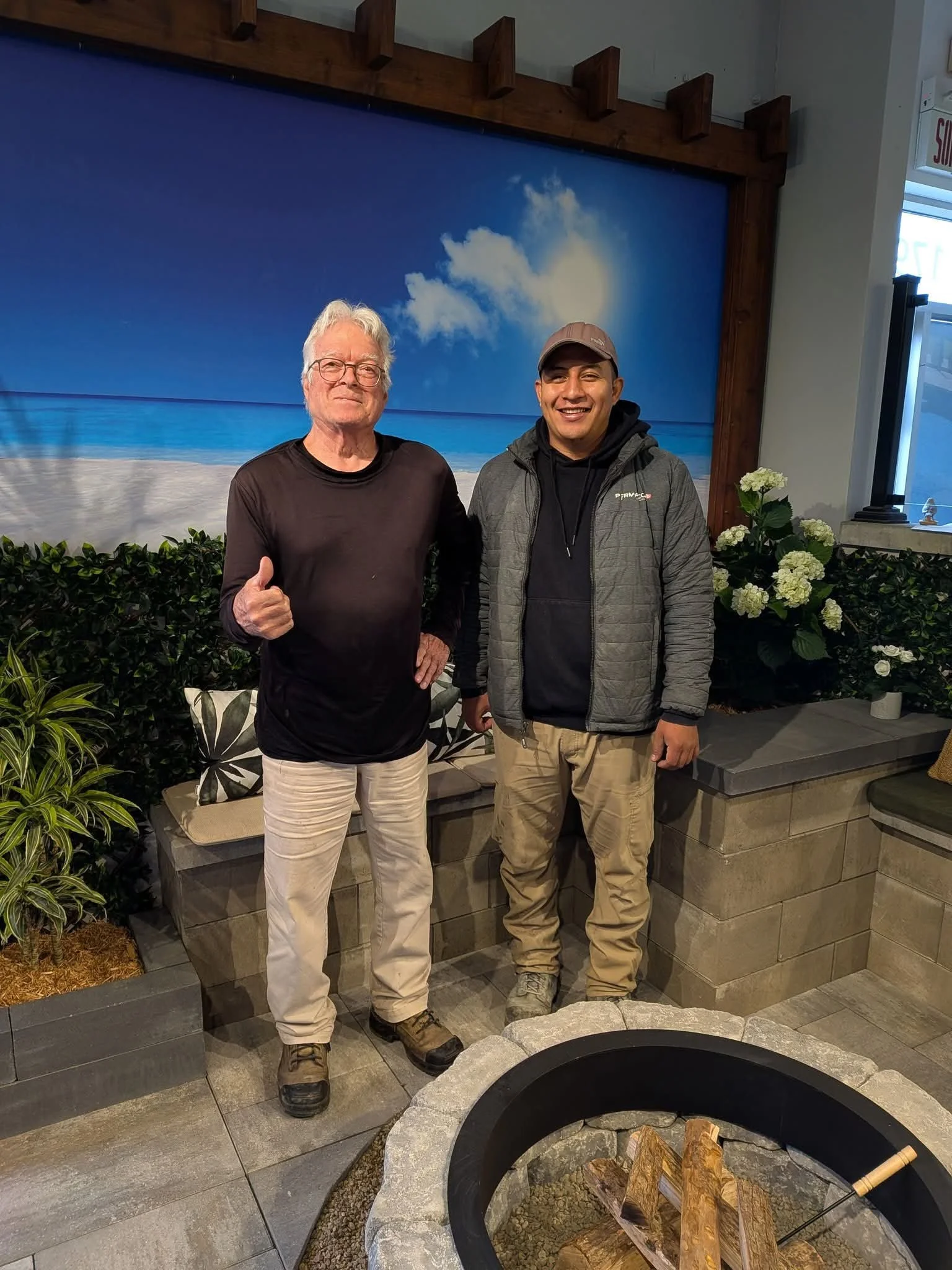 Deux hommes posent côte à côte à l'intérieur d'un restaurant ou café, avec un décor marin en arrière-plan et un foyer rond au premier plan, entouré de plantes et de fleurs, dont des hortensias blanches.