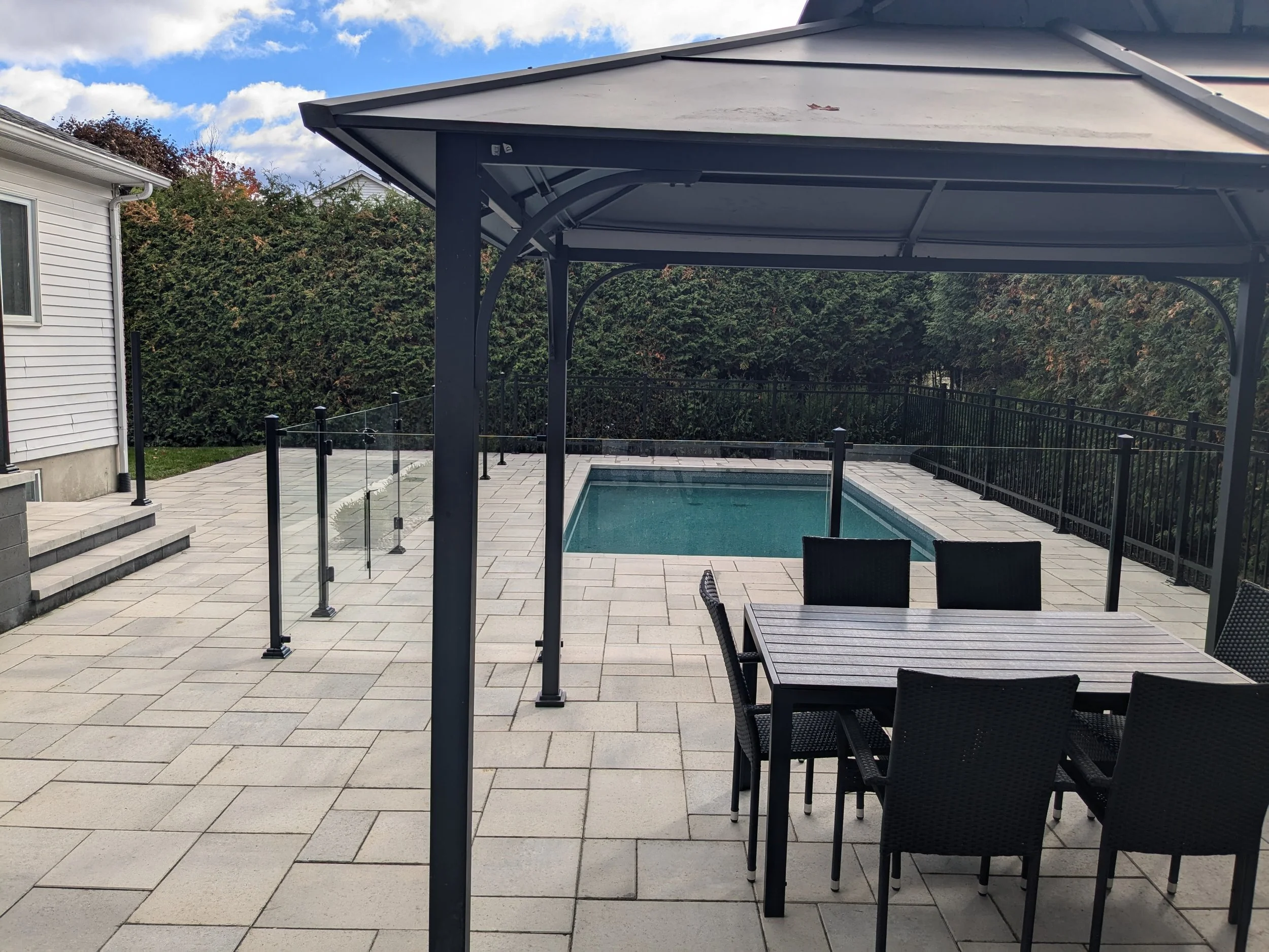Une piscine rectangulaire entourée d'une terrasse pavée, avec un gazebo en métal sombre au premier plan, des chaises en extérieur, une maison à gauche et une haie d'arbustes en arrière-plan.