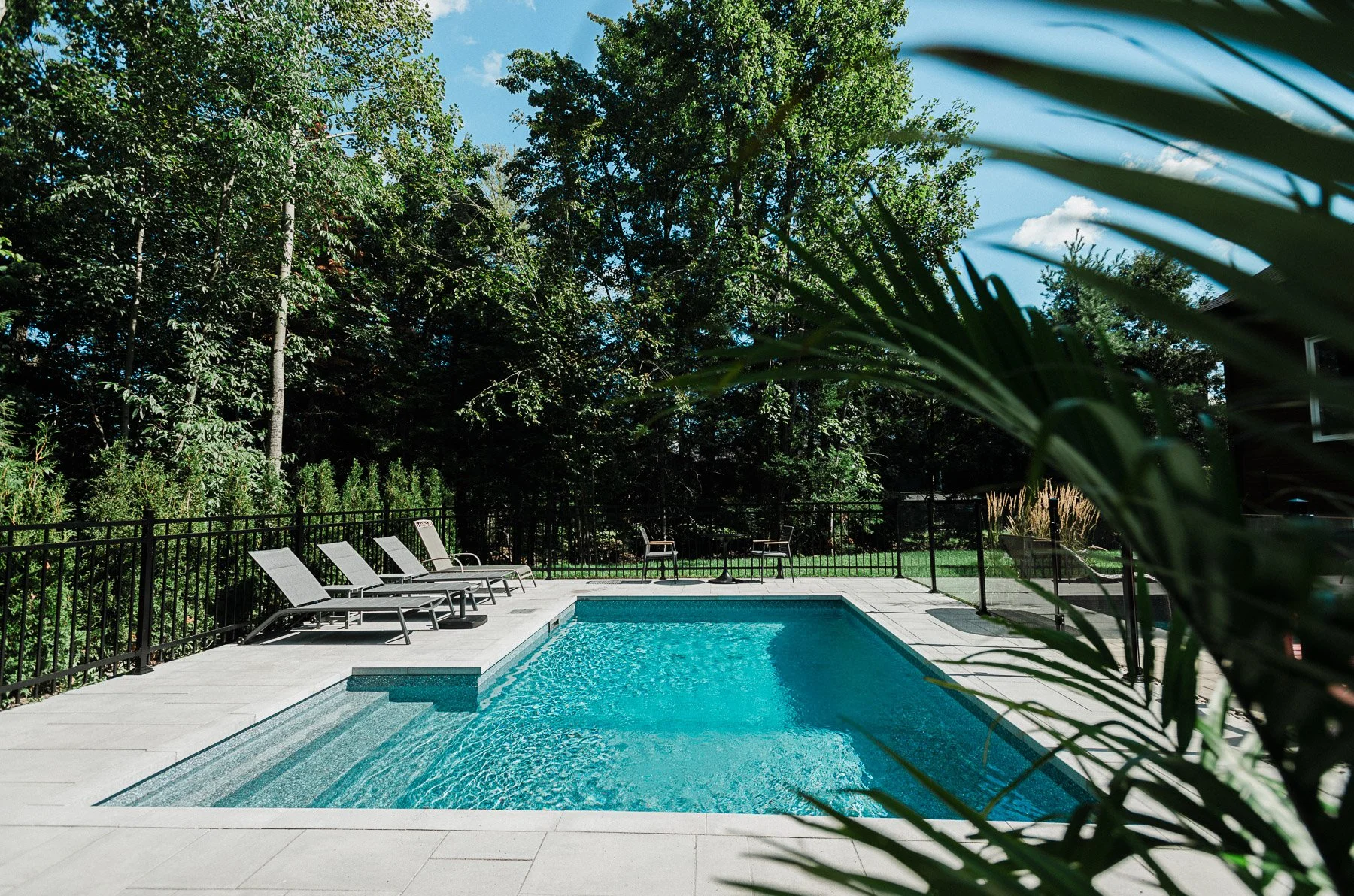 Piscine extérieure entourée d'une terrasse en pierre, avec des chaises longues et des fauteuils, clôturée par une barrière noire, avec un espace vert et des arbres en arrière-plan.