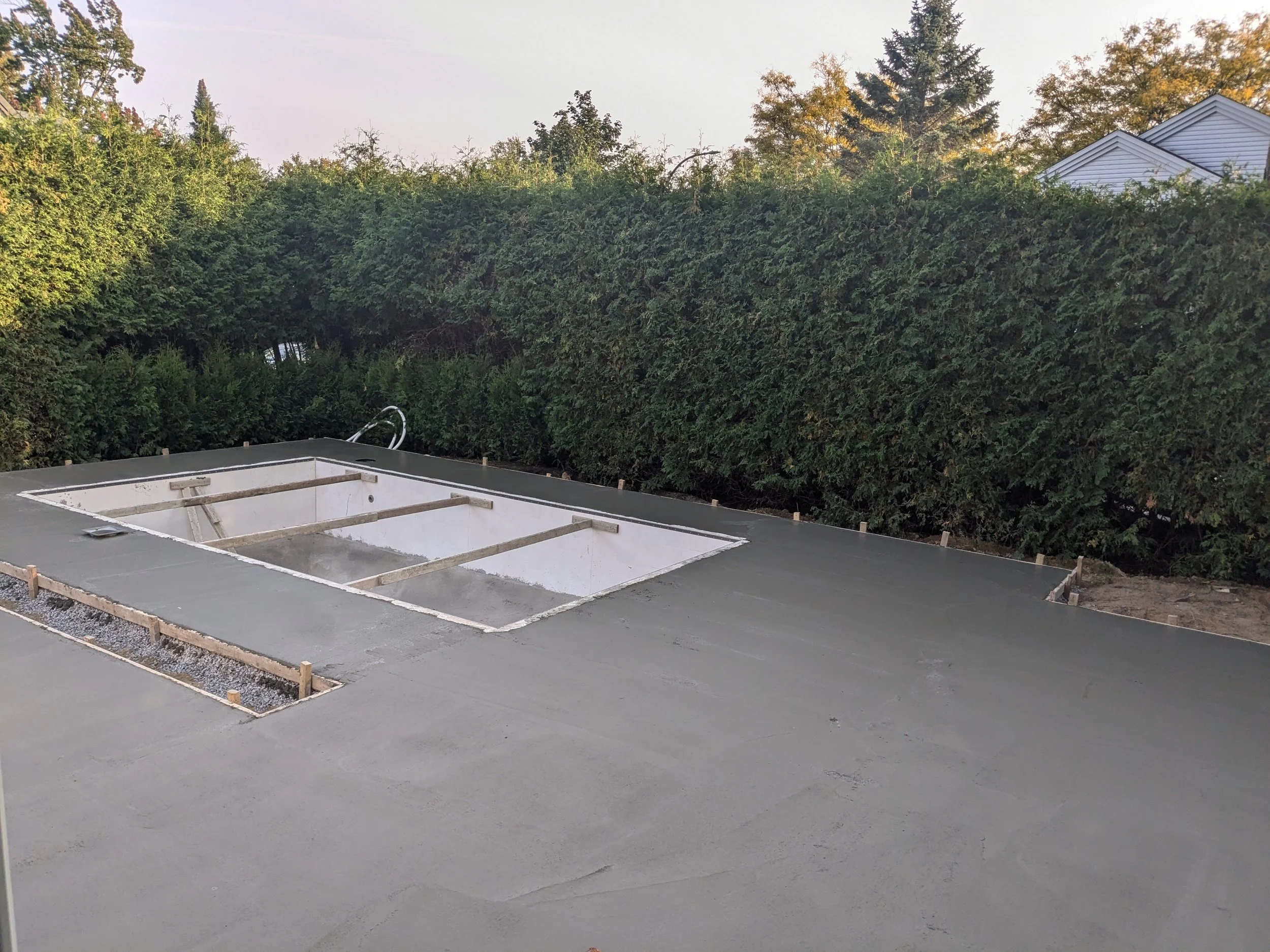 Impression d'une piscine en construction avec une nouvelle couche de béton, entourée de petits piquets en bois pour la délimitation, avec des arbres en arrière-plan.
