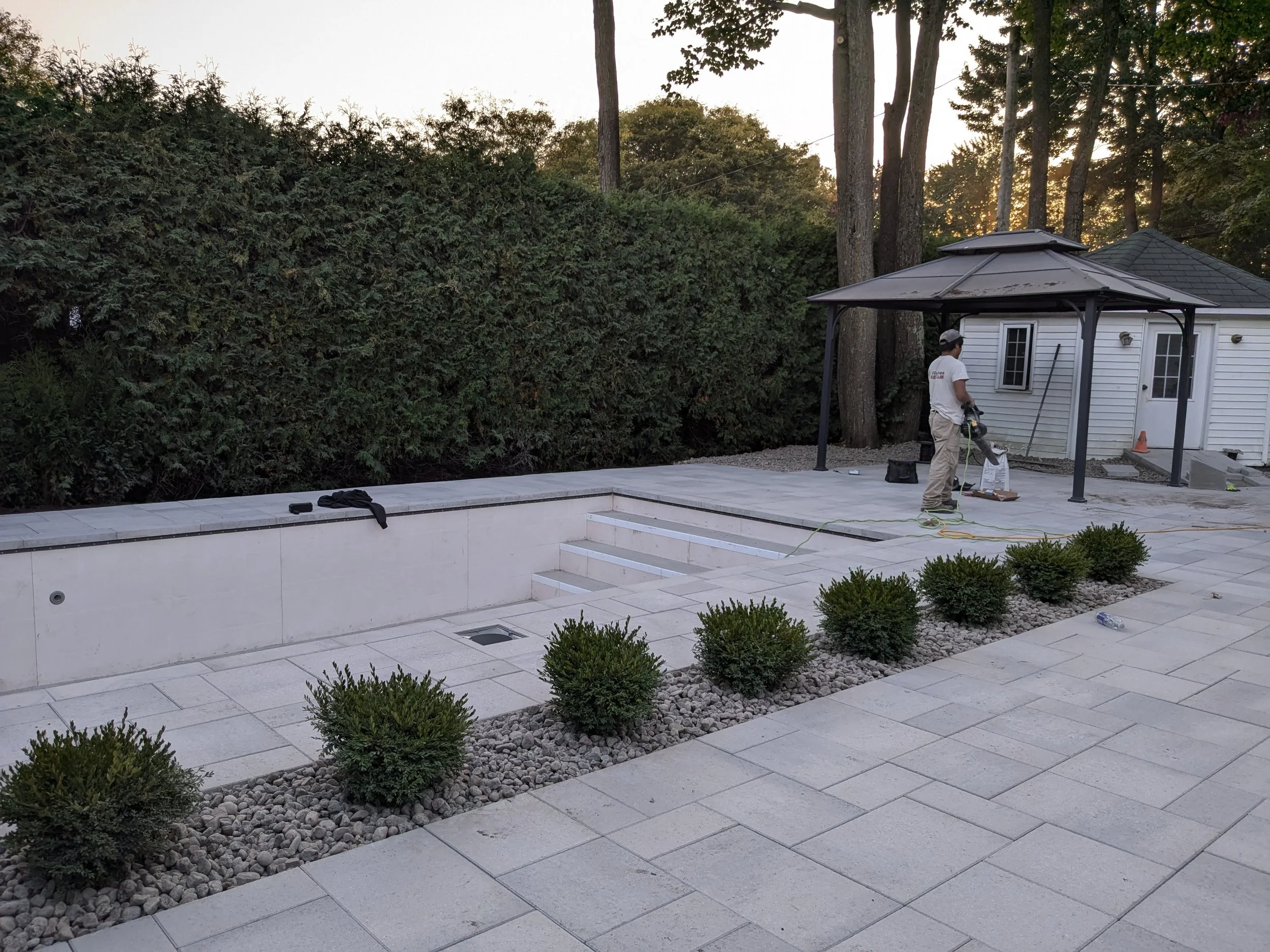 Un homme en train de travailler sur une terrasse en pierre avec une petite maison blanche en arrière-plan, entourée d'arbres et de buissons, à l'extérieur au crépuscule.
