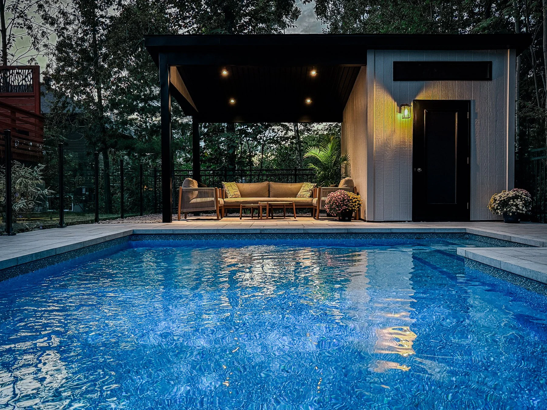 Piscine avec éclairage en soirée, terrasse avec mobilier de jardin, maison moderne en arrière-plan, fleurs décoratives, dans un environnement boisé.