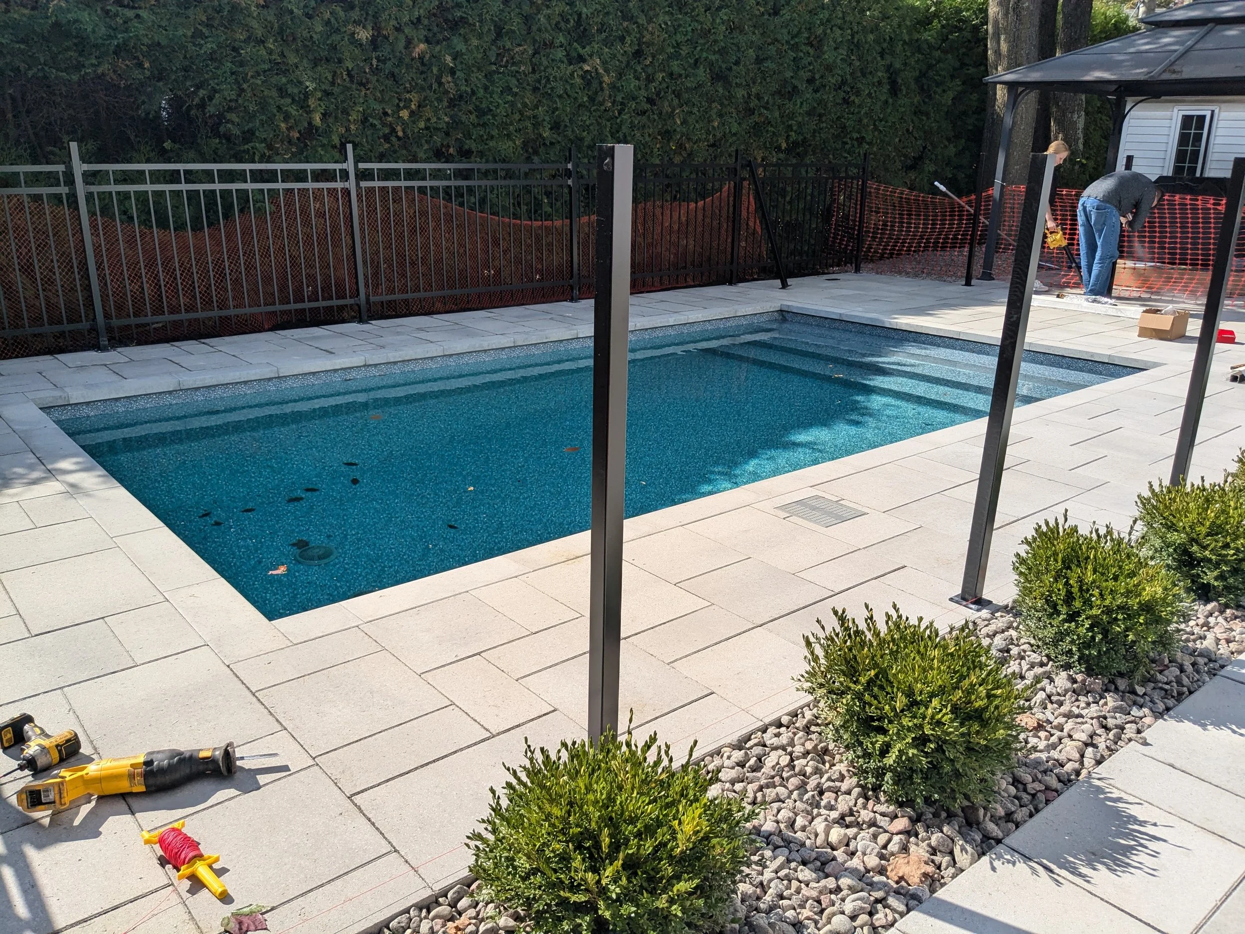 Travaux de construction ou de réparation autour d'une piscine résidentielle, avec des outils électriques posés sur le sol et deux personnes travaillant près d'une clôture en fer noir.