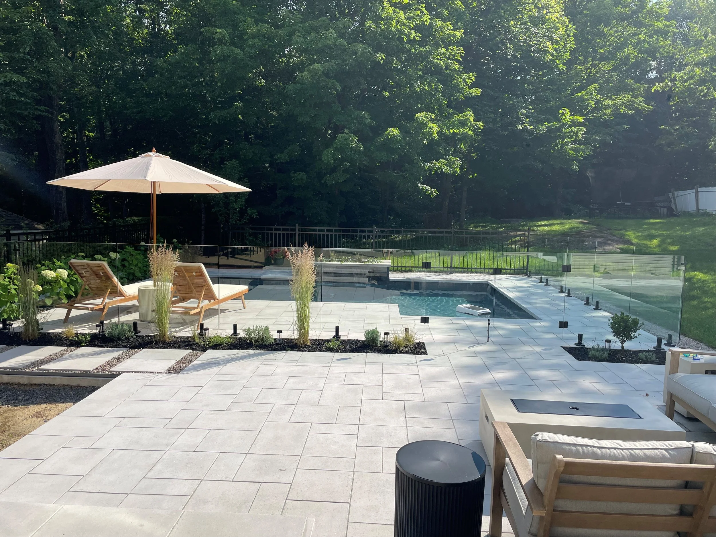 Une piscine extérieure avec des transats en bois et un parasol blanc, entourée d'une terrasse en pierre et d'une barrière en verre, avec un jardin verdoyant et de grands arbres en arrière-plan.