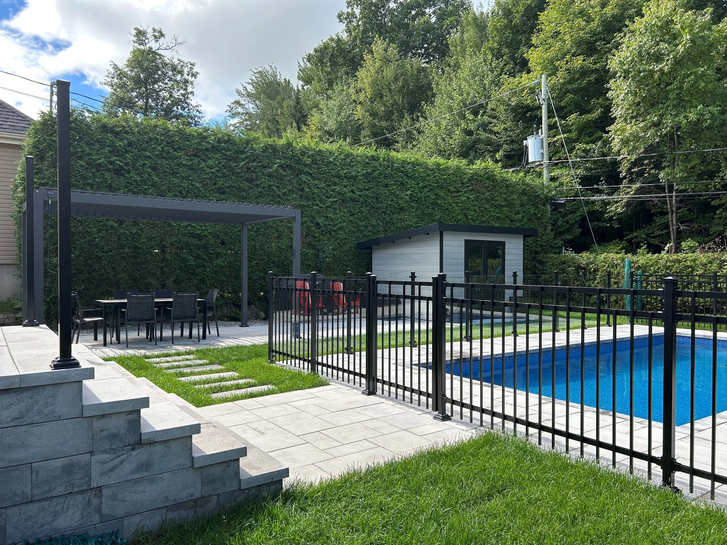 Une piscine bleue entourée d'une clôture noire, avec une zone grillagée et une petite cabane en bois. Il y a un espace de repas avec une grande table et des chaises sous un auvent en métal noir. La scène est située dans un jardin avec des arbres vert