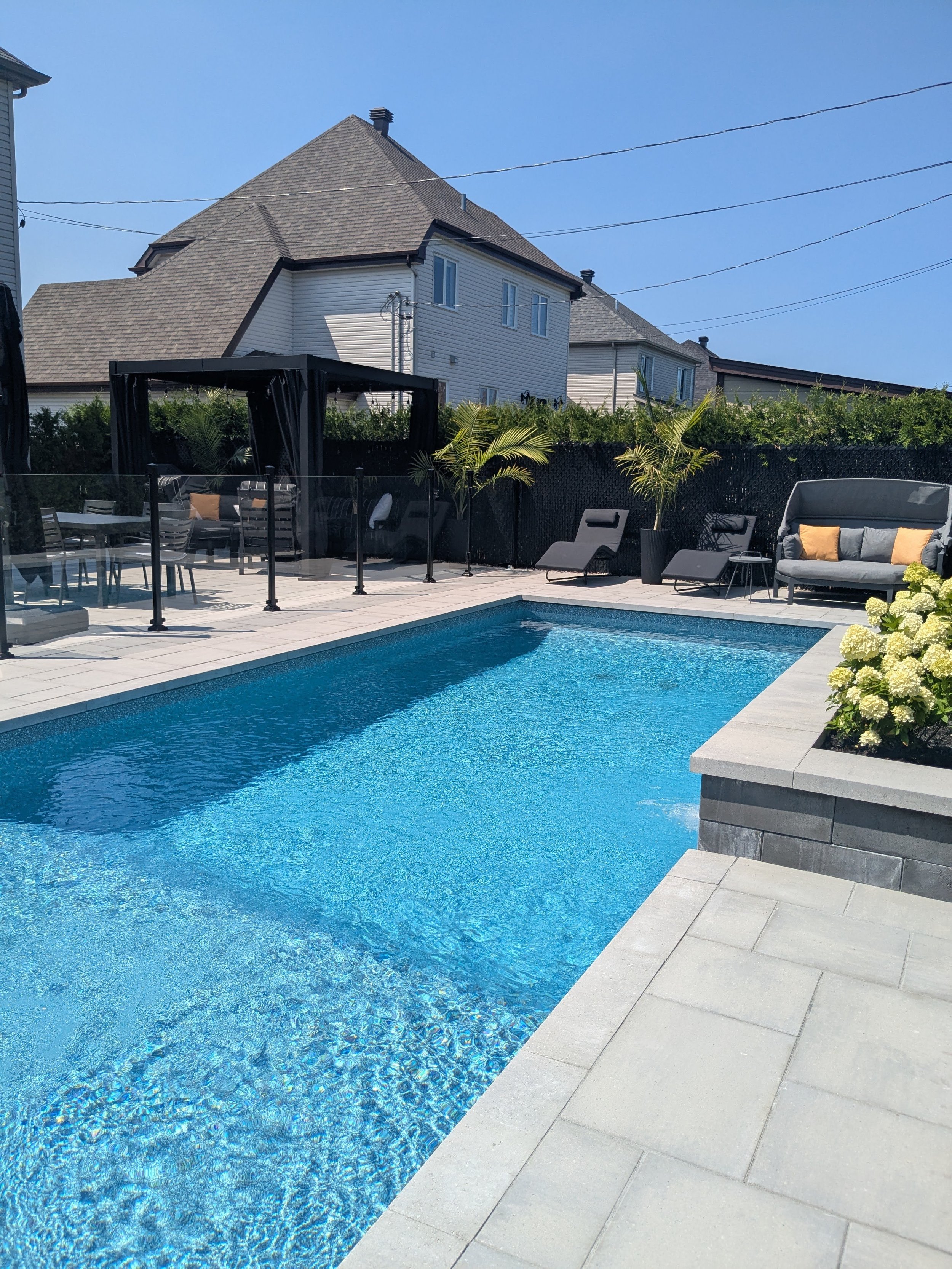 Une piscine extérieure entourée de mobilier de jardin, avec des plantes tropicales, un bâtiment résidentiel en arrière-plan et un ciel dégagé.