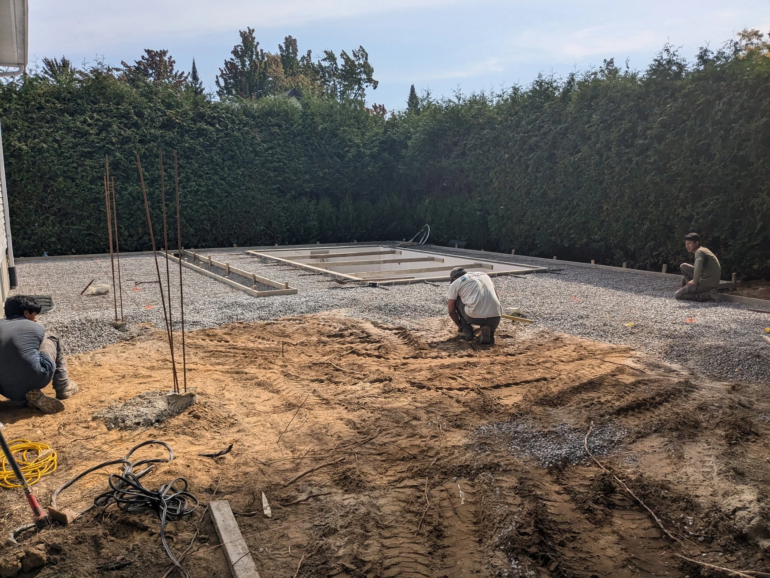 Construction d'une piscine en béton avec outils et ouvriers sur un terrain en plein air entouré d'arbres.