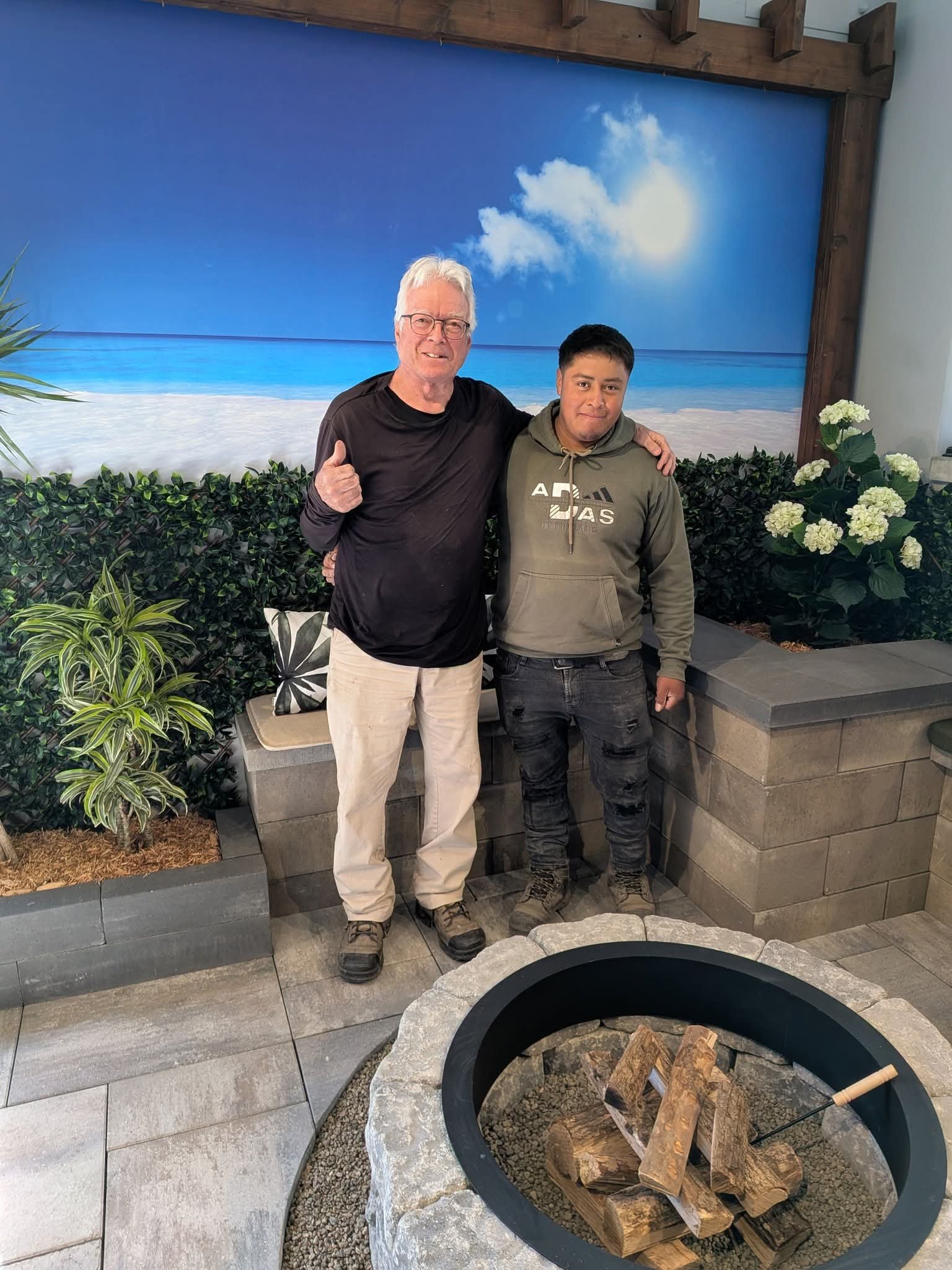 Deux hommes se tiennent côte à côte devant un décor de plage peint, avec des plantes et des fleurs autour, et un feu ouvert contenant du bois en avant.