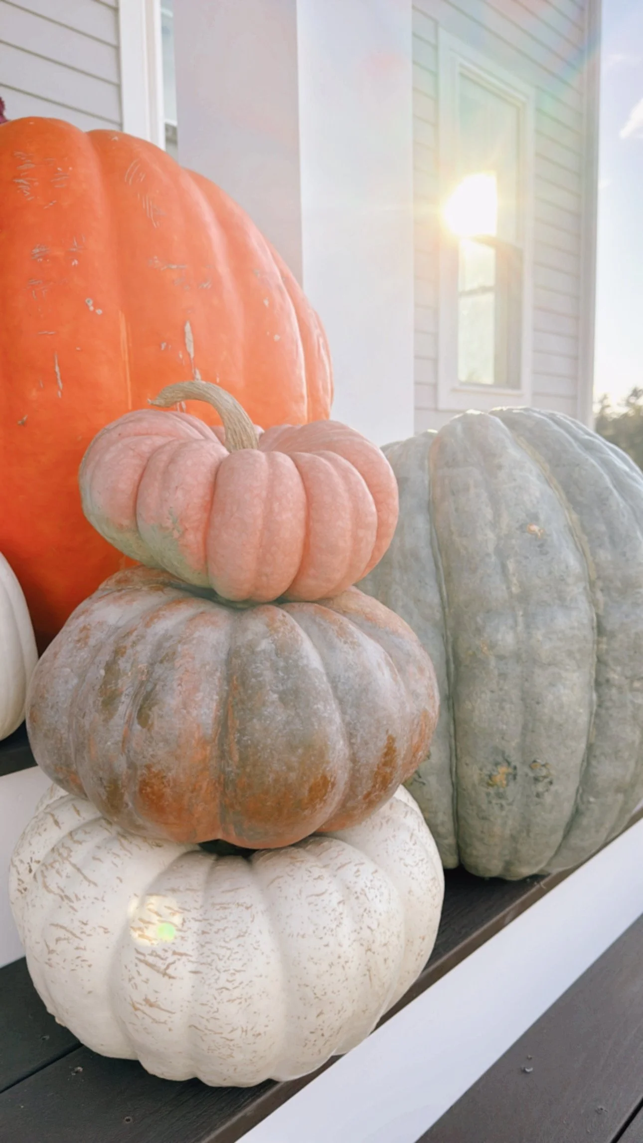 A stack of pumpkins of different sizes and colors, including orange, white, green, and muted pink, arranged on a porch with a house and a window in the background, sunlight shining through.