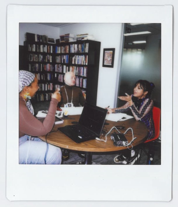 Three women engaged in a discussion around a table with a laptop, recording equipment, and notebooks, in a room with a bookshelf filled with books.