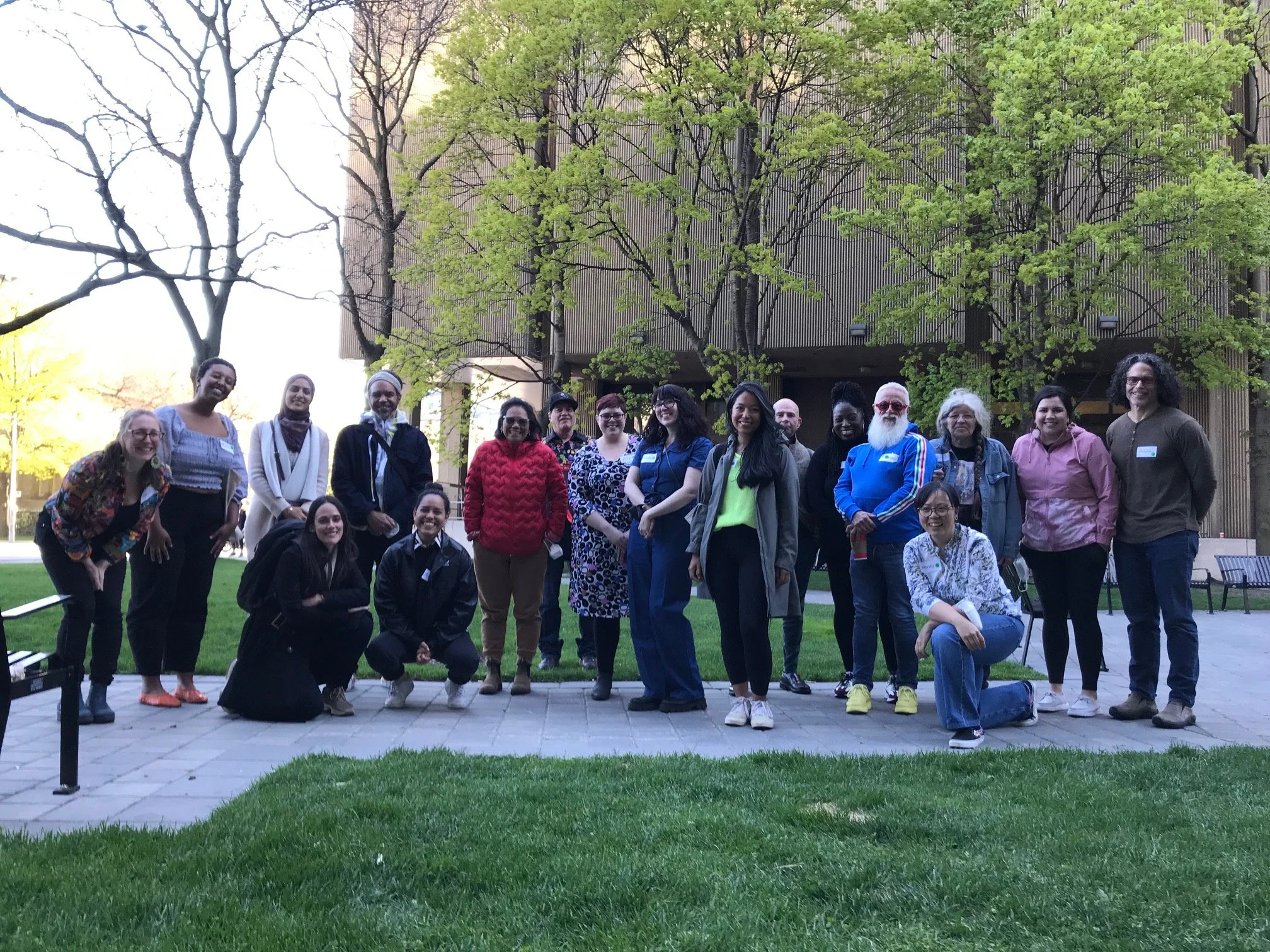 Group of diverse people standing outdoors on a paved area with green grass and trees in the background.