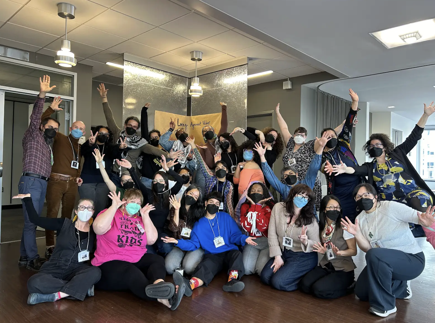 A diverse group of people wearing masks pose together in a modern indoor space, some sitting and some standing, with many raising their hands or making peace signs, celebrating at an event.