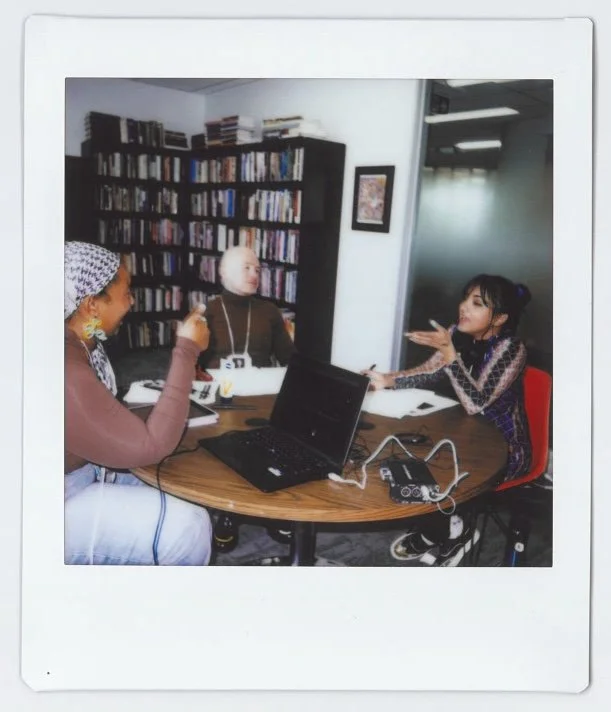 Three women sitting around a round table in a room with a bookshelf filled with books. The woman on the right is talking and gesturing with her hands, while the other two women listen. A laptop and audio equipment are on the table.
