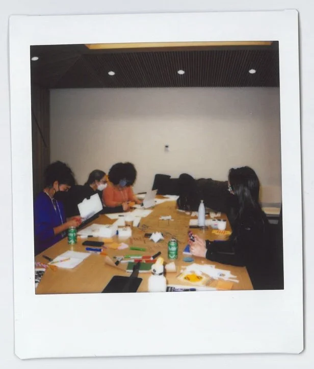 Group of five people wearing masks sitting around a table with papers, markers, and drinks in a conference room.