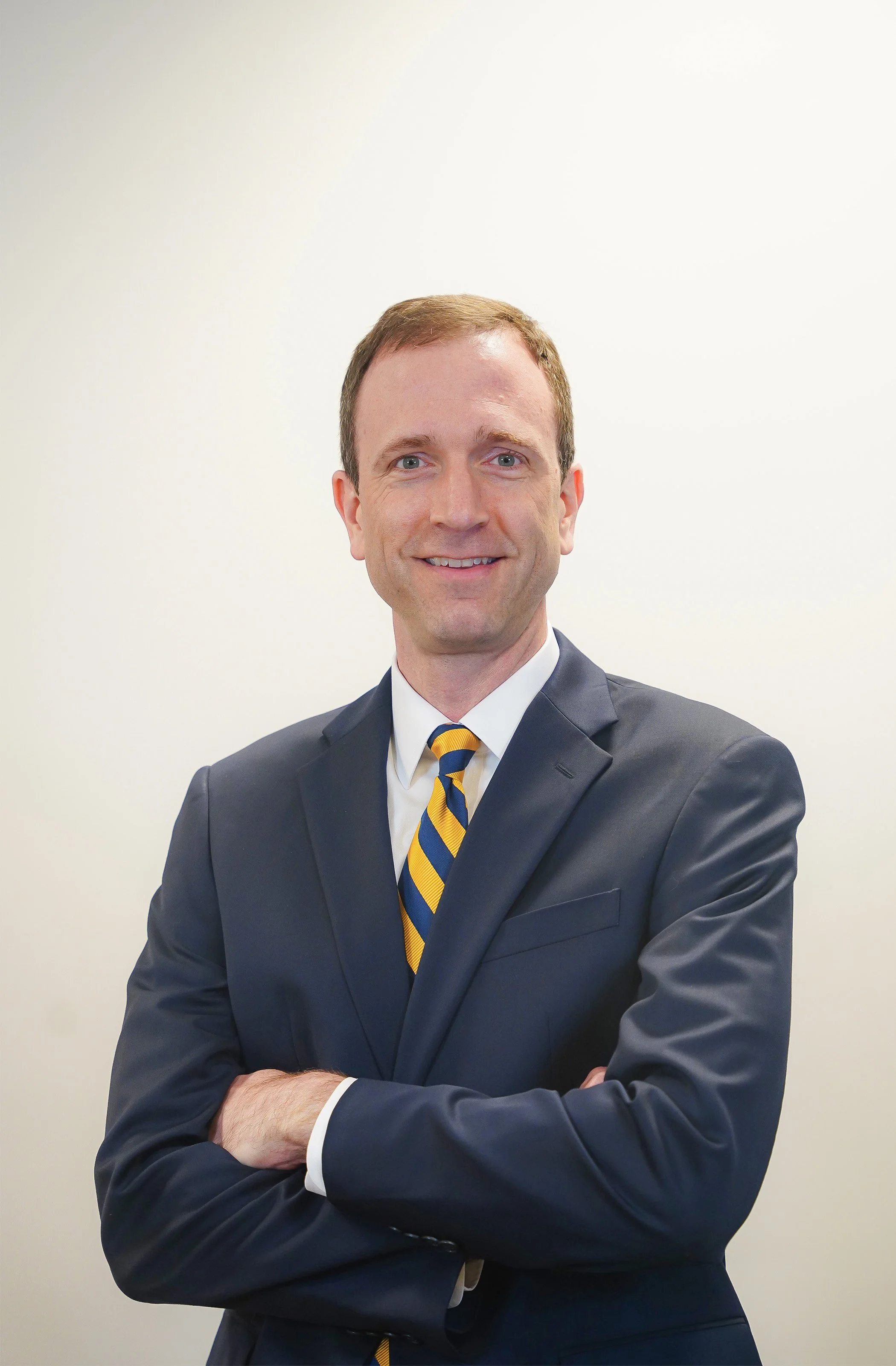 A man in a dark suit with a white shirt and a blue and yellow striped tie crosses his arms and smiles at the camera against a plain white background.