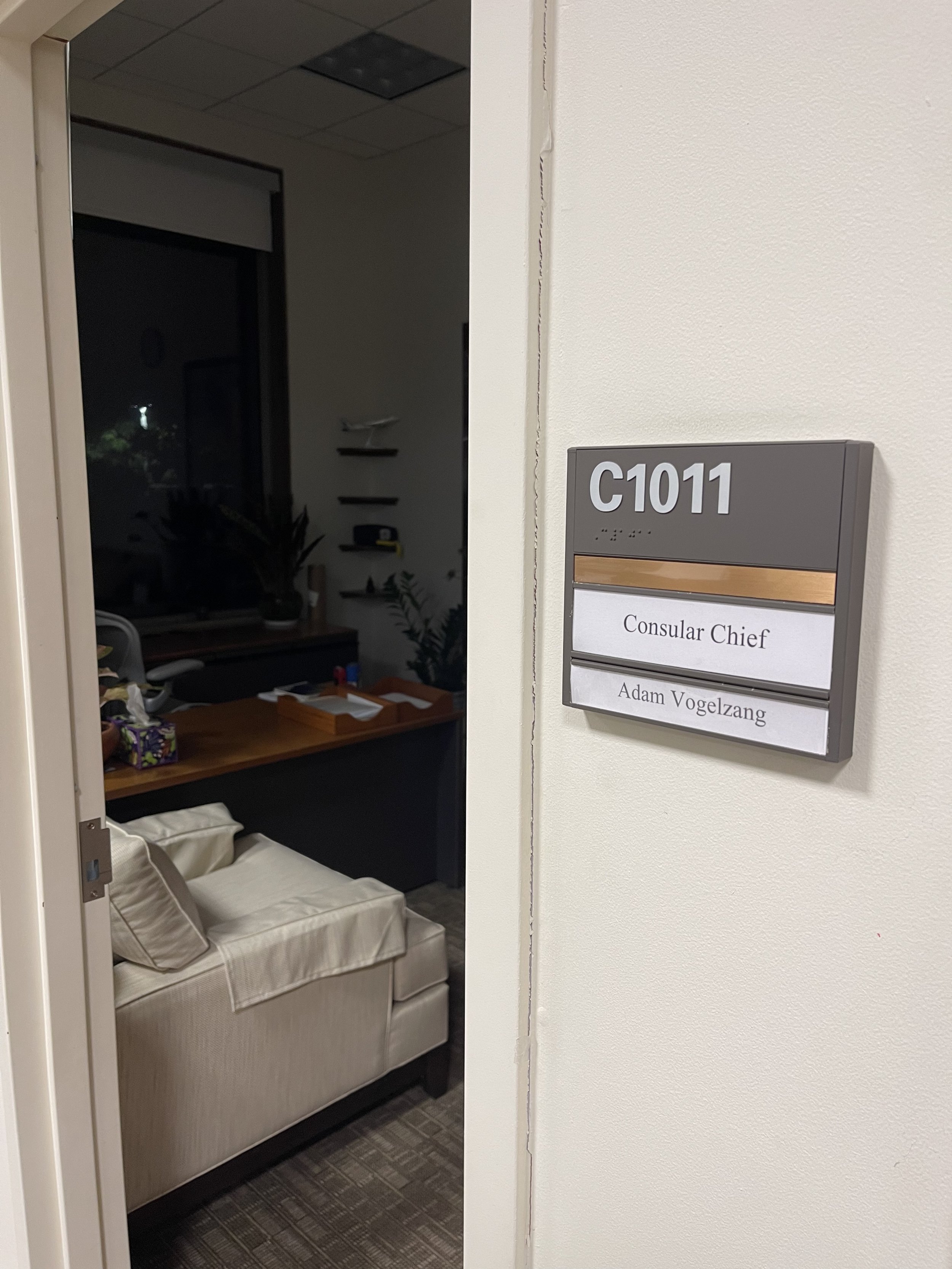 A view inside a room with a beige sofa and pillows, as seen through an open door. There are some black shelves and items on the wall. A window with a dark blind is visible. The wall on the right has a sign that reads 'C1011' with the names 'Consular Chief' and 'Adam Vogelzang' printed below.