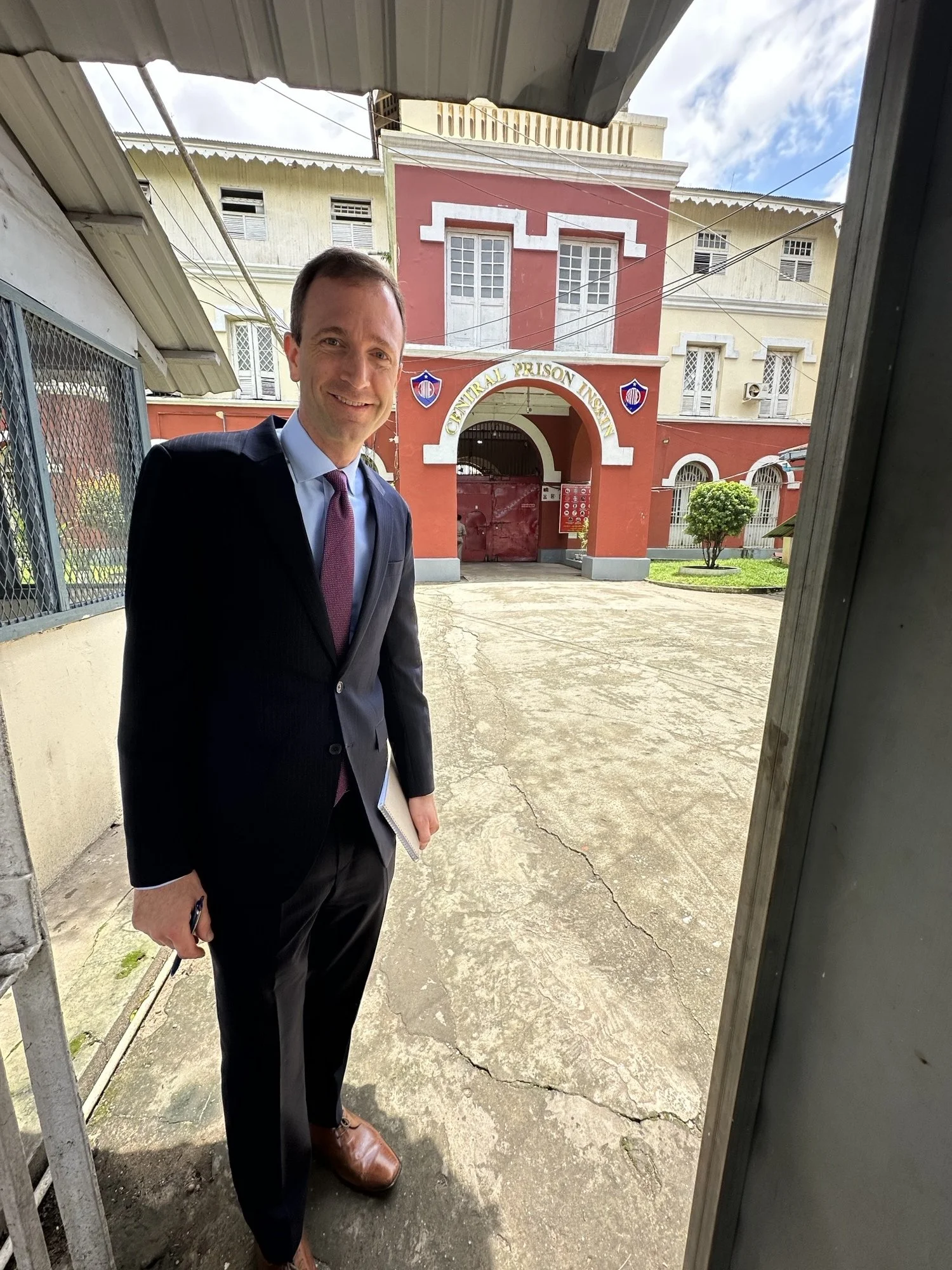 A man in a suit smiling at the camera while standing outside the entrance of the Insein Central Prison Institution building in Yangon, Myanmar.