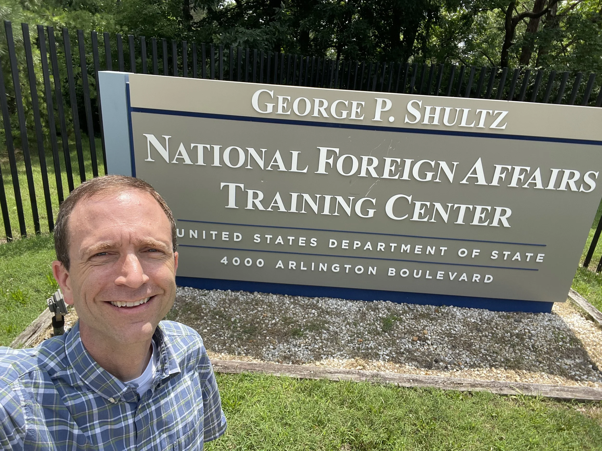 A man smiling, wearing a plaid shirt, taking a selfie in front of a sign at the George P. Shultz National Foreign Affairs Training Center with trees and grass in the background.