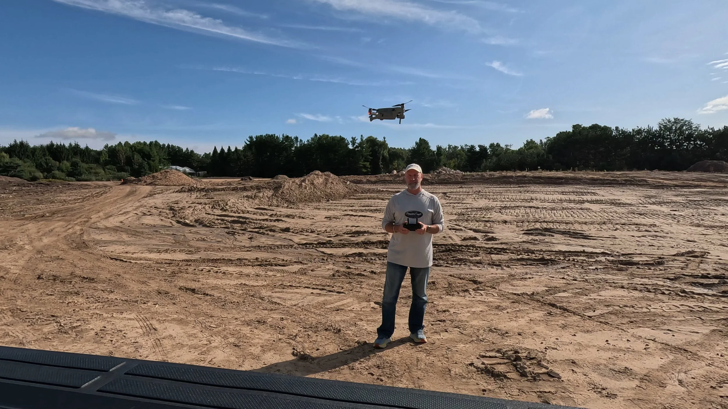 A man standing in a dirt lot holding a remote control, with a drone flying in the sky above him, nearby trees, blue sky, and tire tracks in the dirt.