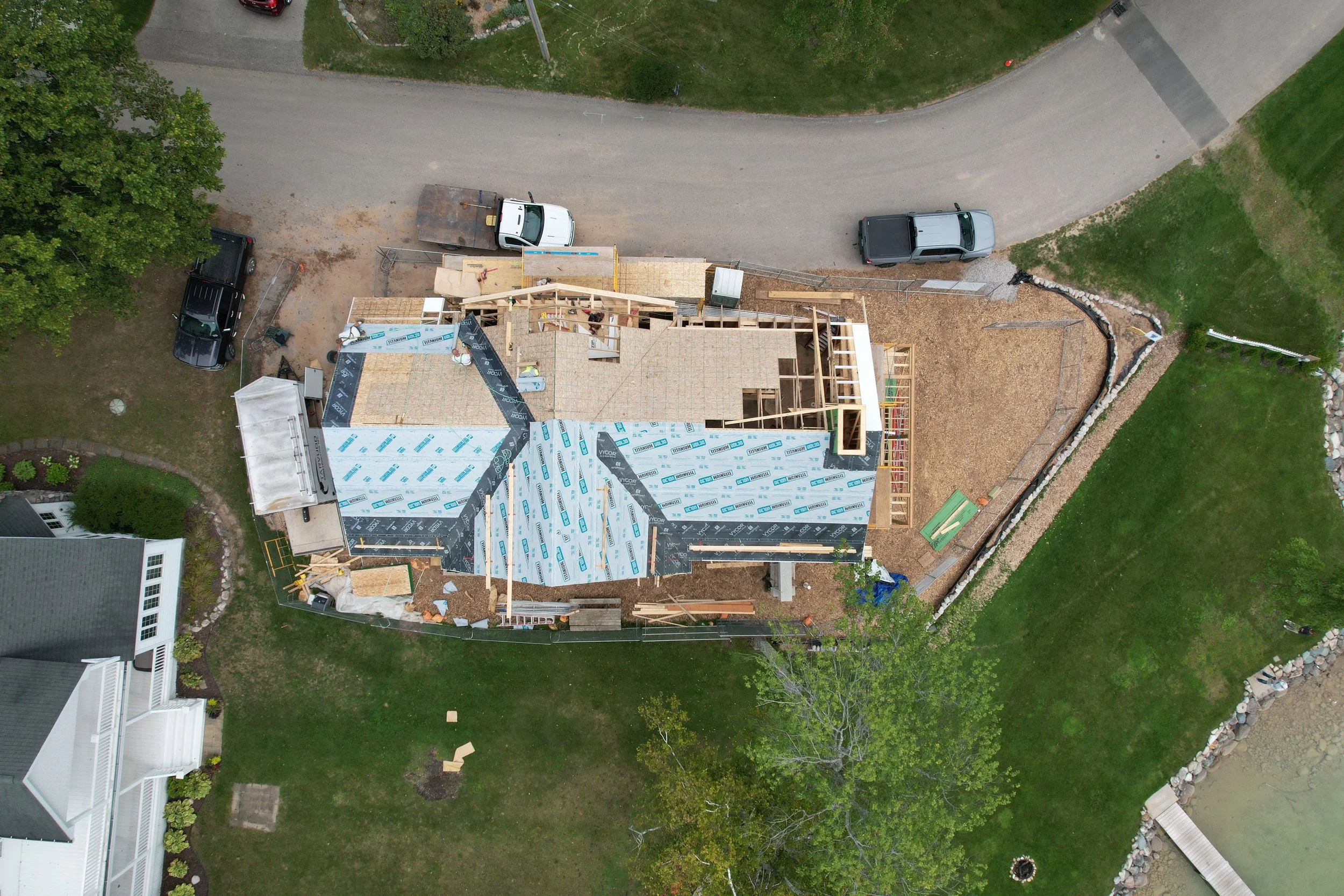 An aerial view of a house under construction with framing and roofing in progress, surrounded by a green lawn, trees, and a gravel driveway with three parked vehicles.