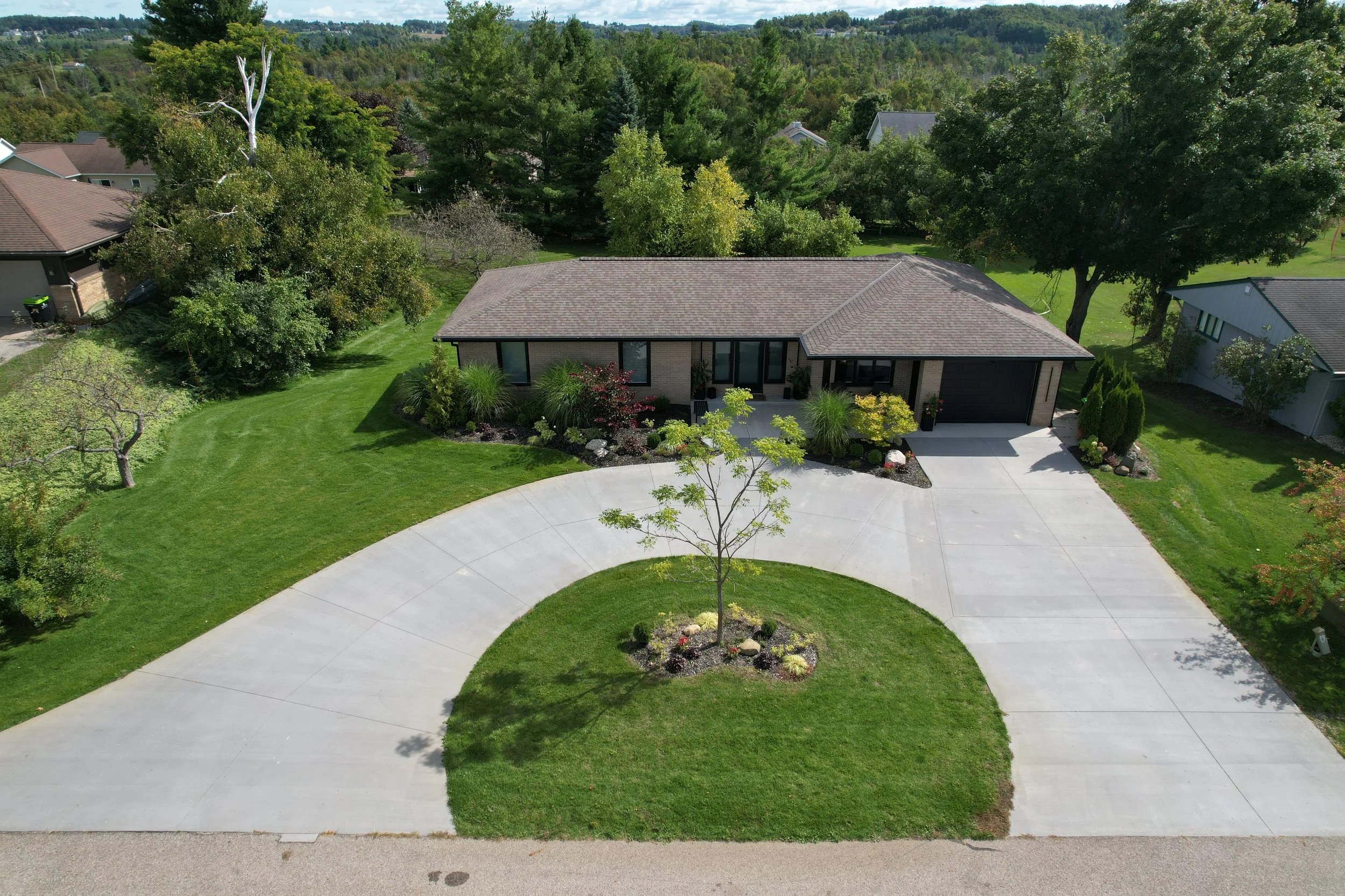 Aerial view of a modern single-story house with a gray roof, surrounded by well-maintained green lawn and landscaped garden with bushes and flowering plants. The house has large windows and a garage. The driveway curves around the front yard. Tall trees and neighboring houses are in the background, with a wooded hillside in the distance.