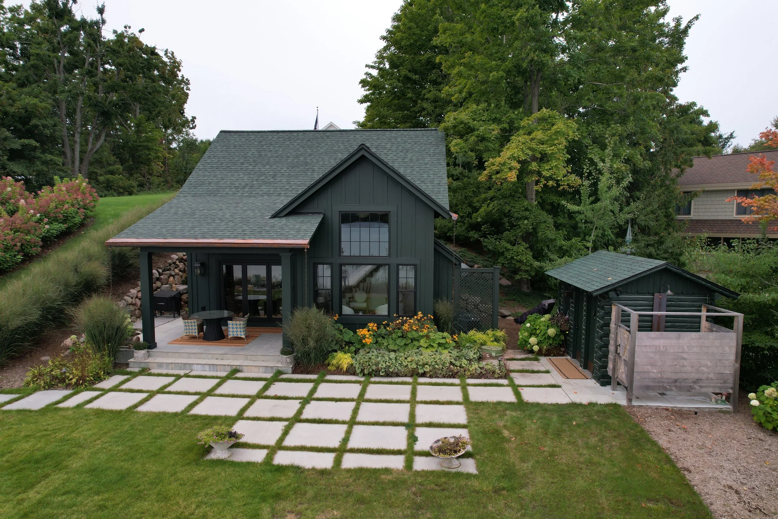A modern dark gray house with large windows and a gabled roof, surrounded by lush greenery, trees, and a well-maintained garden with flowering plants. There is a patio area with outdoor furniture and a stone pathway leading to the house.