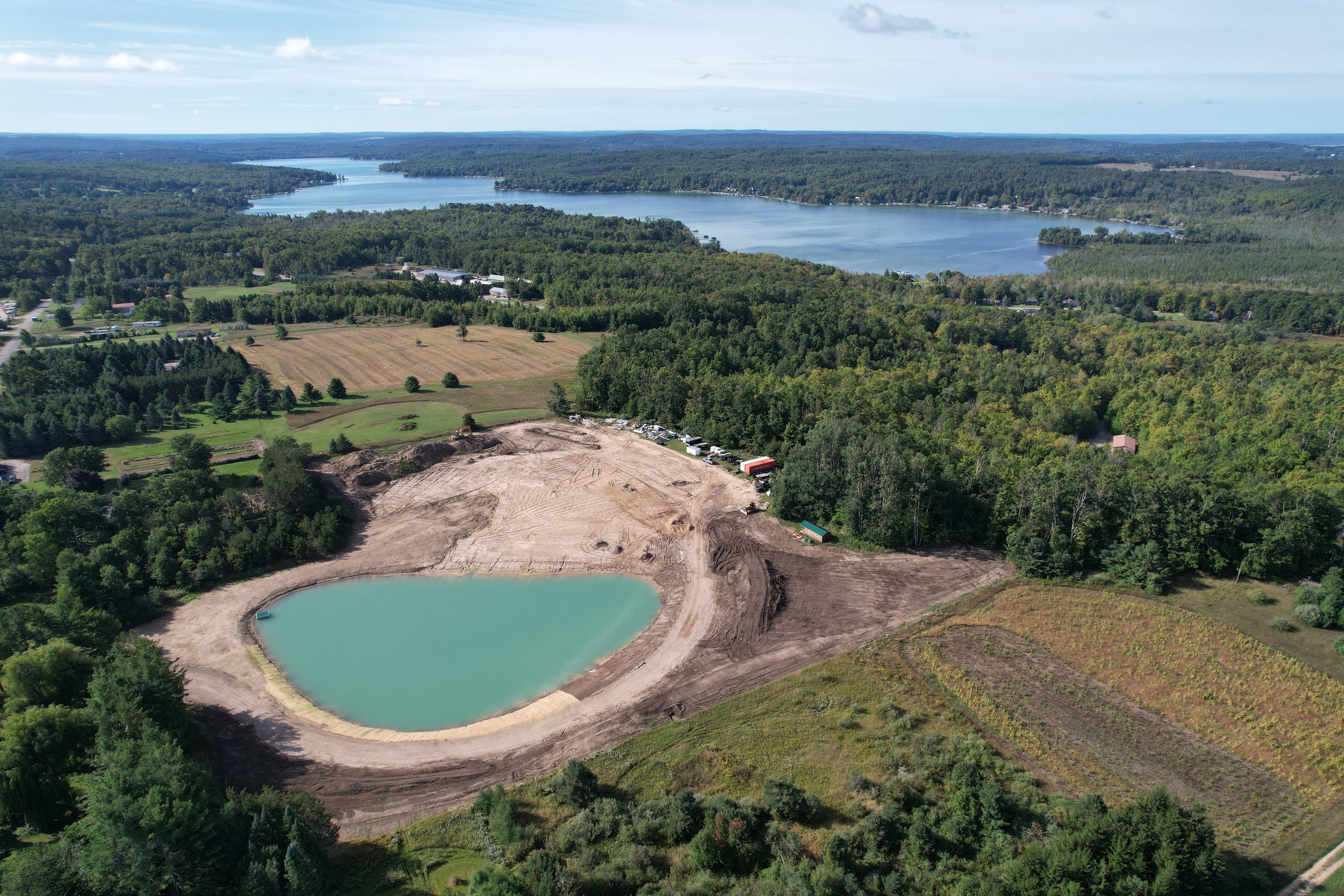 Aerial view of a large body of water surrounded by dense green forests, with some development and residential areas along the shoreline and an area under construction with dirt and a small artificial pond.